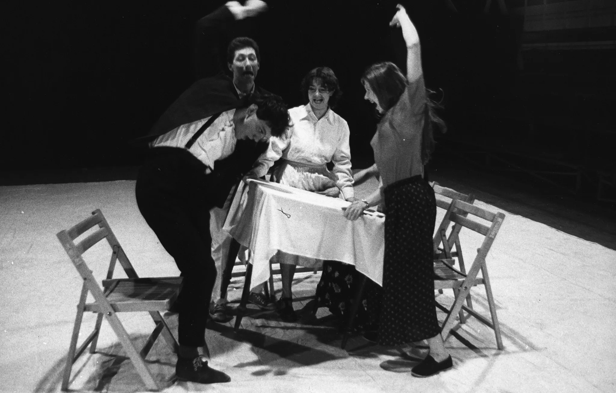 Przy stole stoi pięcioosobowa grupa aktorów i aktorek. Wszyscy wyglądają na zdenerwowanych. Niektórzy unoszą stół, inni w niego uderzają.