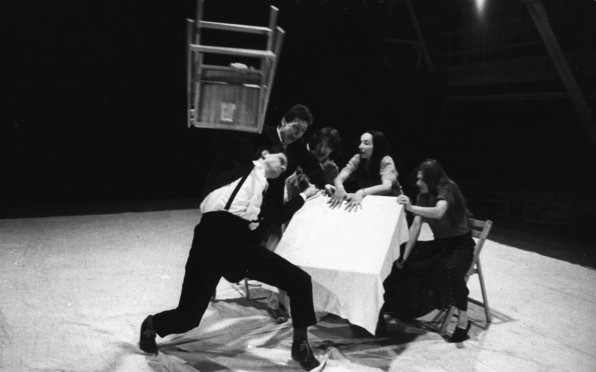 Pięcioosobowa grupa aktorów i aktorek przy stole. Wyglądają na zdenerwowanych. Stół jest przechylony. Aktor z lewej strony trzyma wysoko w górze krzesło.