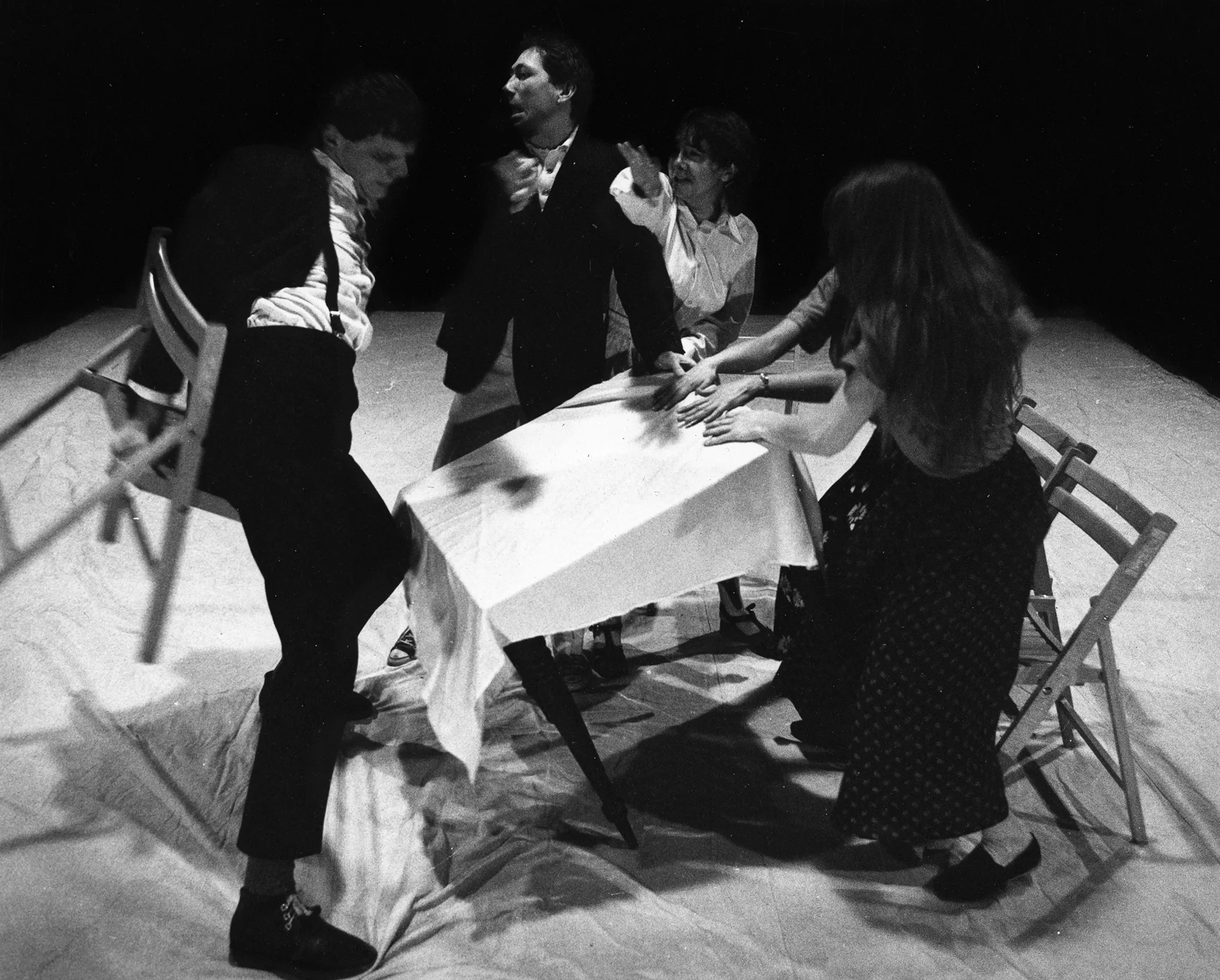 Grupa aktorów i aktorek przy stoliku. Wyglądają na zdenerwowanych. Stolik jest przechlony. Aktor po lewej stronie unosi krzesło.