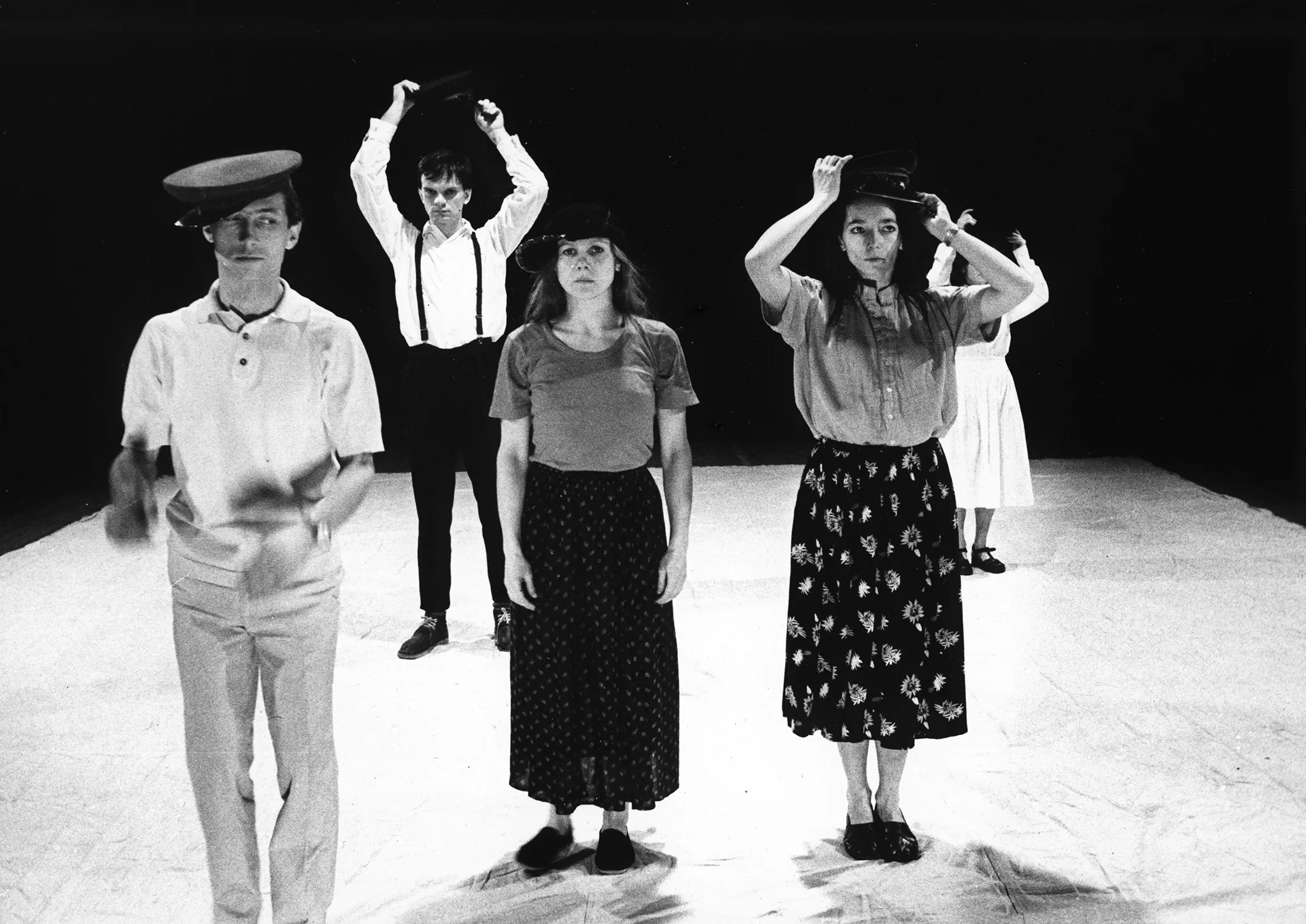Na białej płachcie stoi pięcioosoba grupa aktorów i aktorek zwróconcych twarzą w stronę widza. Wyglądają, jakby byli ubrani prywatne. Wzrok przykuwają ich duże nakrycia głowy.