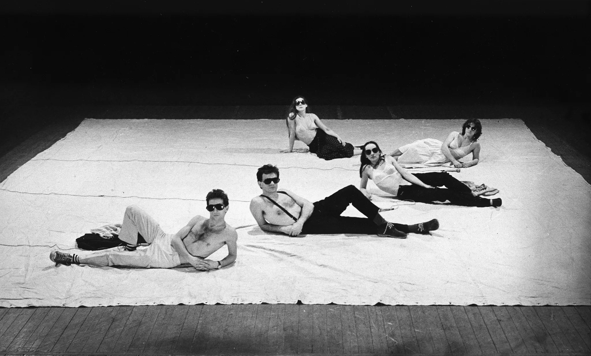 Pięcioosobowa grupa aktorów i aktorek leży na białej płachcie. Wyglądają, jakby pozowali do zdjęcia. Aktorzy mają nagie torsy, aktorki mają na sobie staniki. Wszyscy noszą ciemne okulary przeciwsłoneczne.