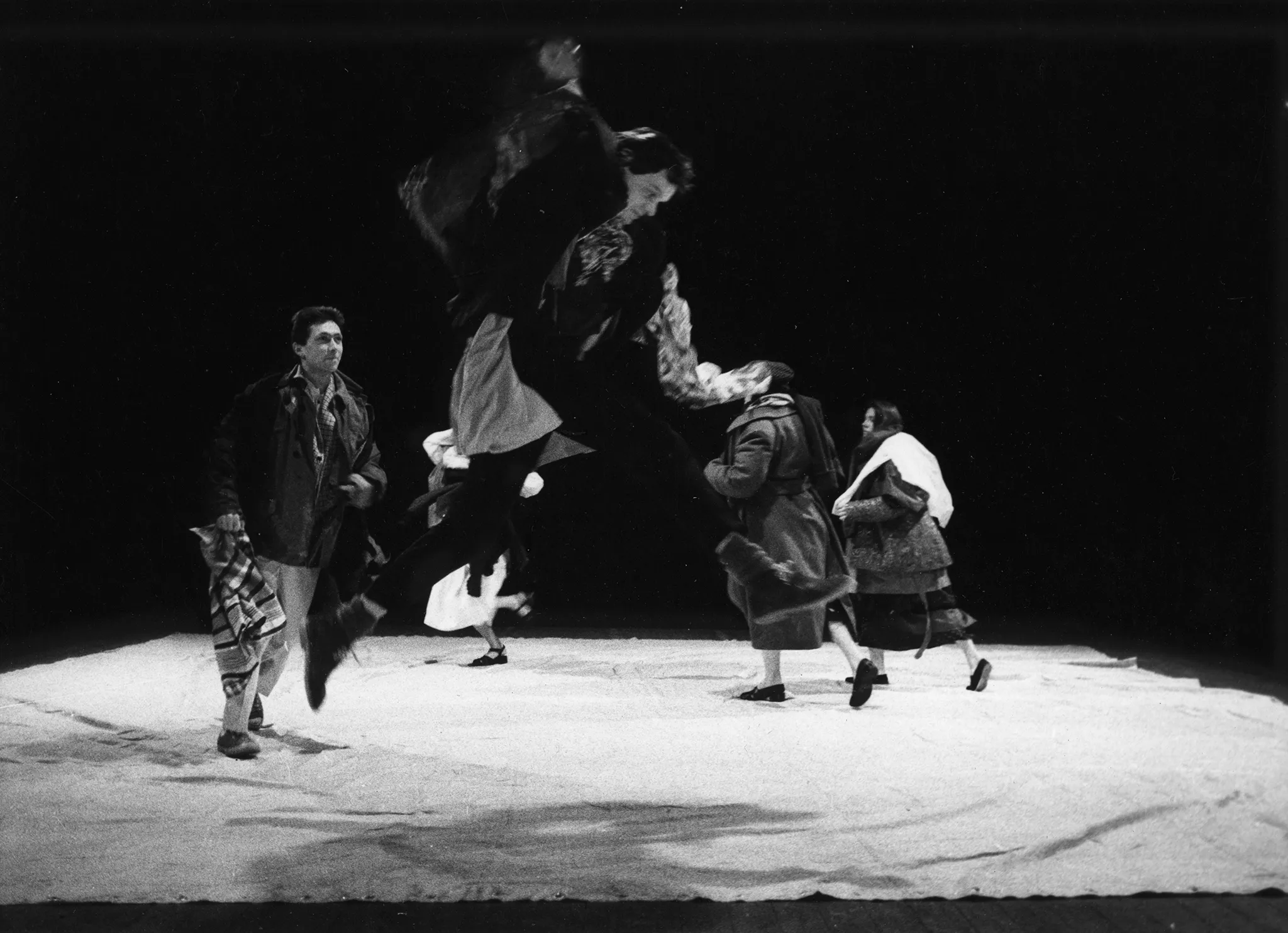 Grupa aktorów i aktorek na białej płachcie. Wyglądają, jakby mieli na sobie zbyt dużo ubrań. Wykonują ekspresyjne ruchy. Na pierwszym planie aktor wykonuje bardzo wysoki skok.