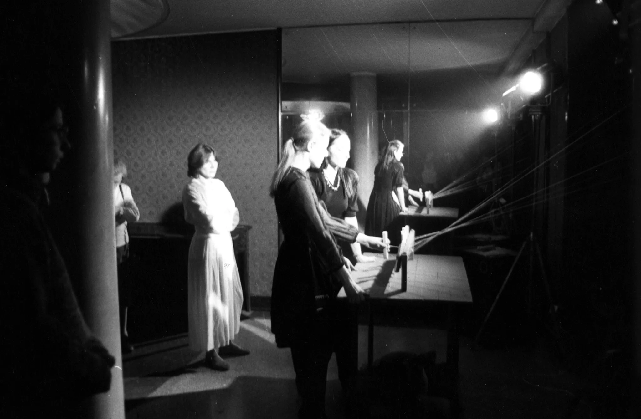 Niewyraźne zdjęcie. W pomieszczeniu stoi grupa aktorek zwróconych profilem w stronę widza. Dwie aktorki stoją rzy stoliku. Na stoliku ustawiony jest trudny do zidentyfikowania przedmiot złożony z drewnianych elementów i długich linek. Na tylnej ścianie wisi duże lustro, w którym wszystko się odbija.