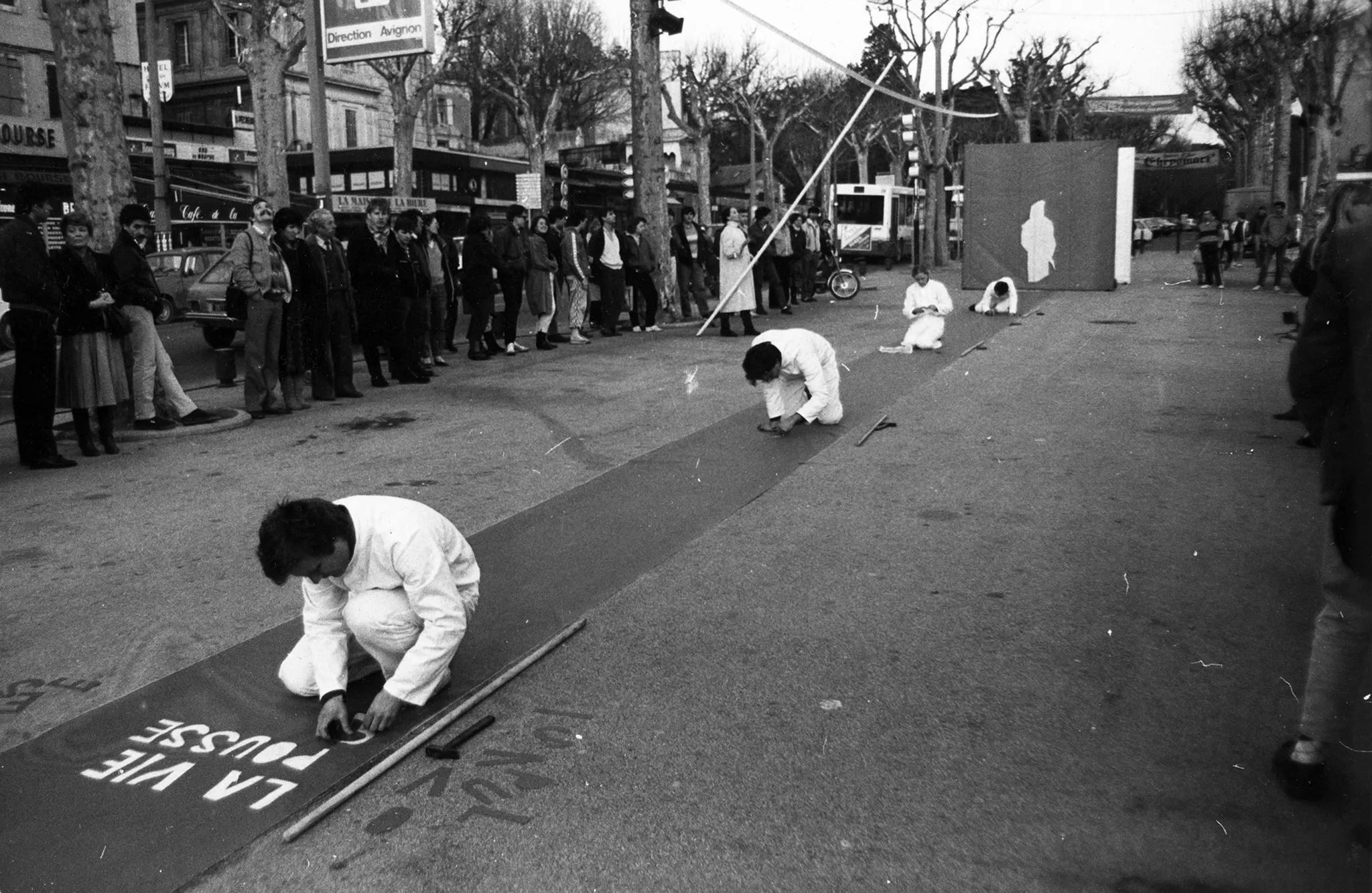 Przestrzeń miejska. Na środku ulicy rozwinięty wybieg. Na wybiegu siedzi czteroosobowa grupa aktorów i aktorek ustawionych jedno za drugim w dużej odległości. Ubrani są na biało. Przyklejają do wybiegu białe napisy. Po dwóch stronach stoi przyglądająca im się grupa widzów. W tle widać drzewa, budynki i samochody.