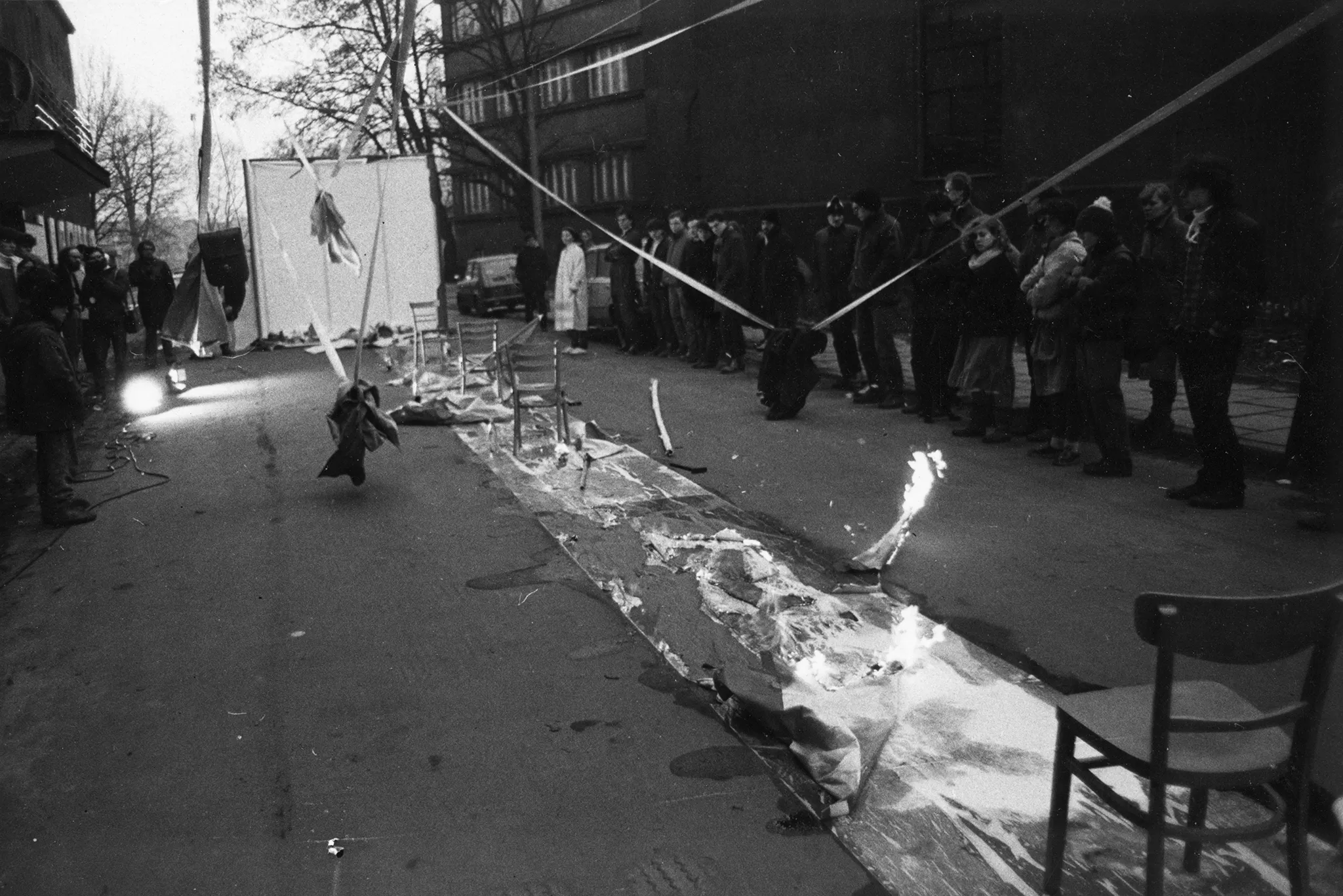 Przestrzeń miejska. Na środku ulicy rozwinięty wybieg. Wybieg jest mokry. Stoją na nim krzesła. W niektórych miejsach wybiegu pali się ogień. Po obu stronach wybiegu stoi publiczność. W tle widać budynki, drzewa i samochody.