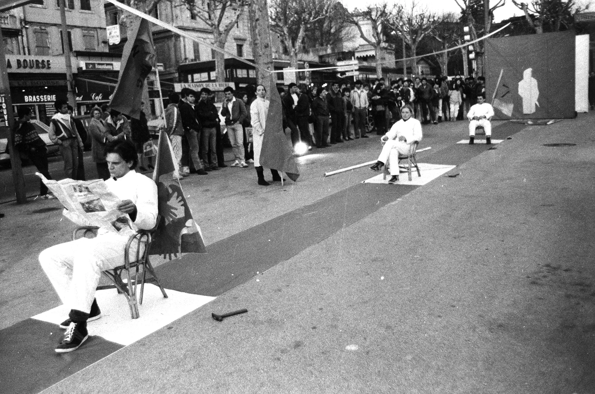 Przestrzeń miejska. Na środku ulicy rozwinięty wybieg. Na wybiegu, na krzesłach siedzi dwóch aktorów i jedna aktorka. Ustawieni są jedno za drugim w dużej odległości. Ubrani są na biało. Aktor siedzący z przodu czyta gazetę. Obok wybiegu stoi grupa widzów. W tle widać drzewa i budynki.