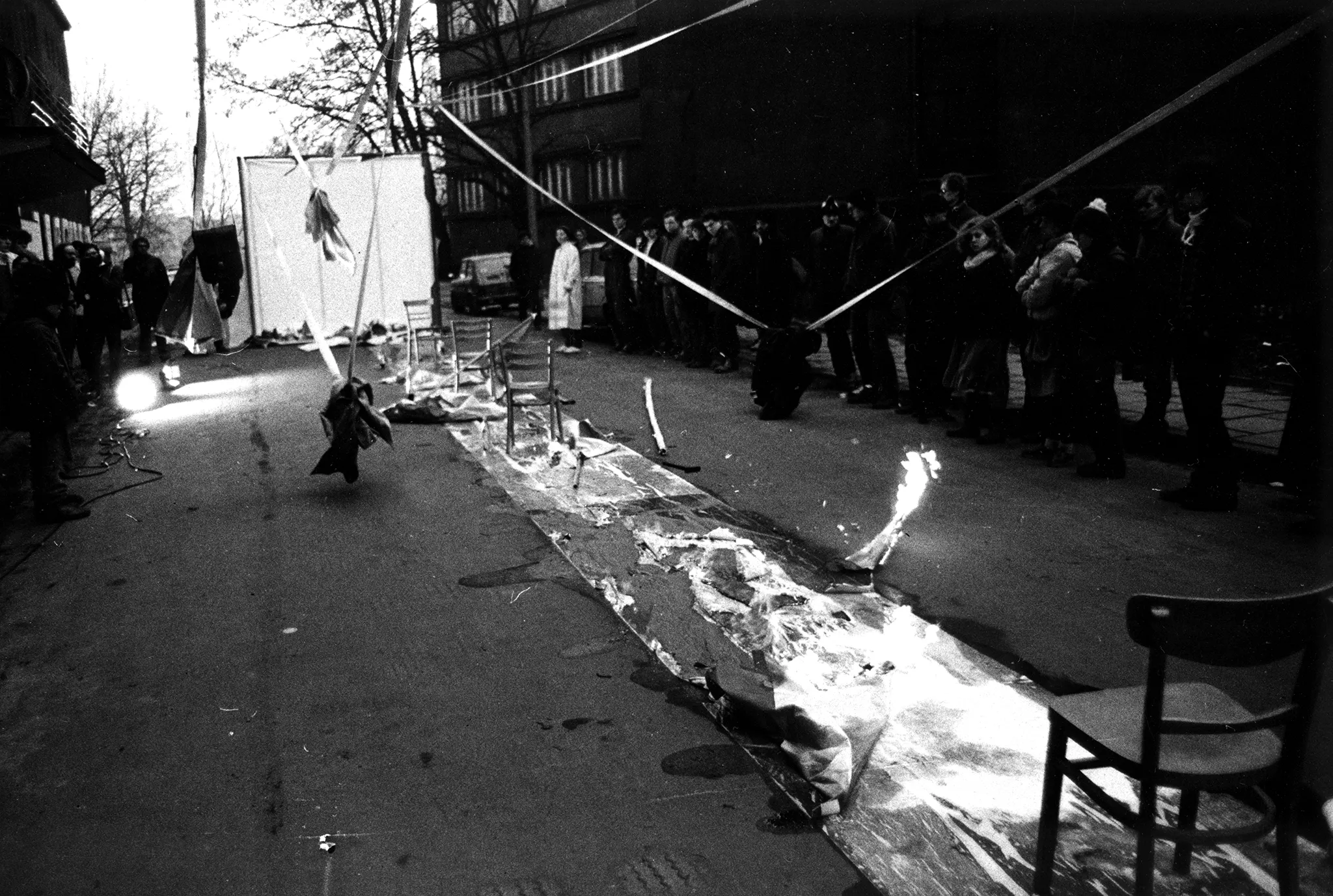 Przestrzeń miejska. Na środku ulicy rozwinięty wybieg. Wybieg jest mokry. Stoją na nim krzesła. W niektórych miejsach wybiegu pali się ogień. Po obu stronach wybiegu stoi publiczność. W tle widać budynki, drzewa i samochody.