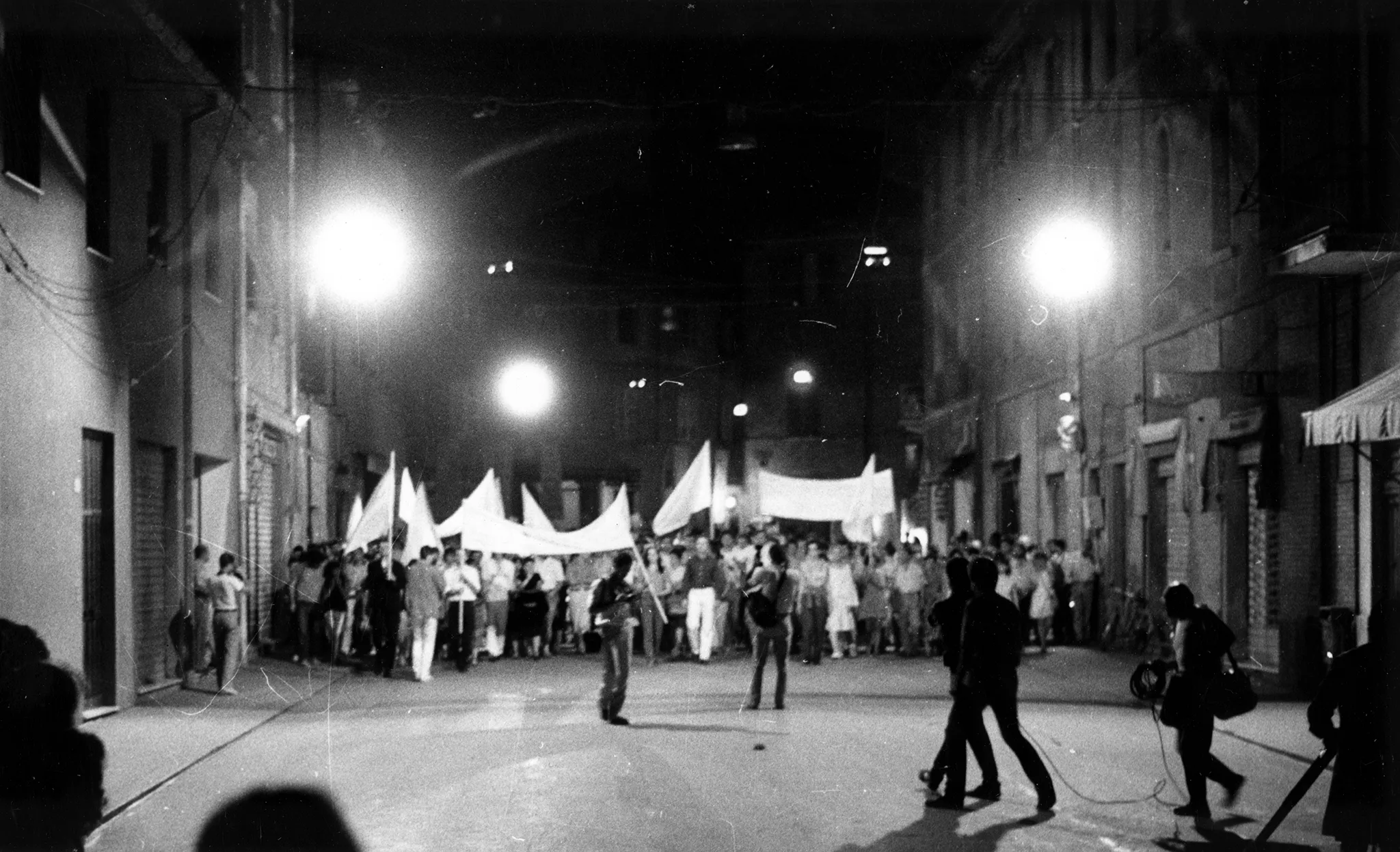 Niewyraźne zdjęcie. Przestrzeń miejska. Między budynkami stoi tłum ludzi. Niektórzy trzymają w rękach białe flagi. W stronę tłumu podchodzi ekipa filmowa.