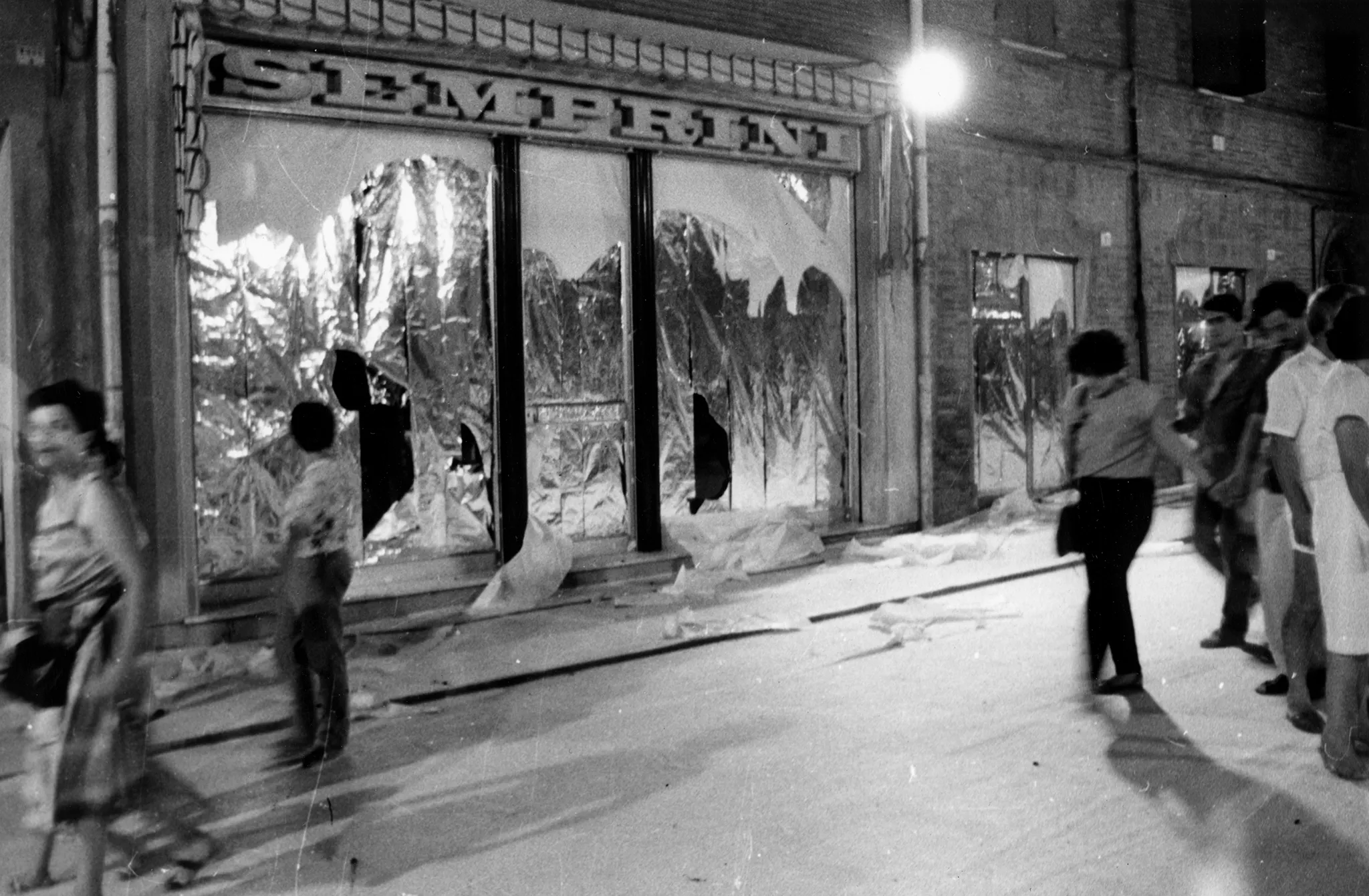 Przestrzeń miejska. Ulica wyglądająca jak tuż po zamieszkach. Pod zasłoniętymi witrynami sklepowymi leżą białe flagi. Ulicą idzie tłum ludzi.