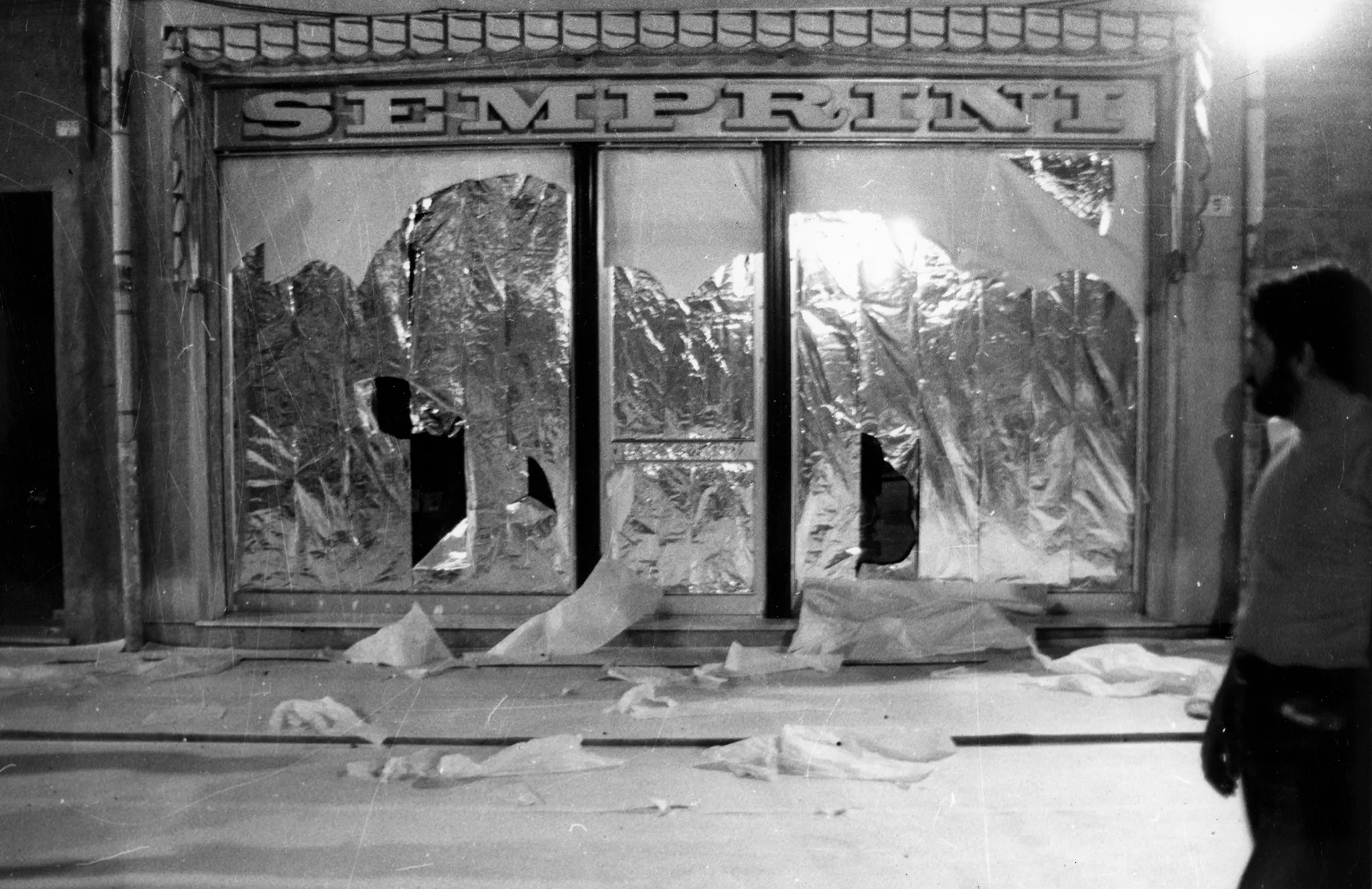 Przestrzeń miejska. Ulica wyglądająca jak tuż po zamieszkach. Pod zasłoniętymi witrynami sklepowymi leżą białe flagi. Nad drzwiami budynki widnieje napis "SEMPRINI".