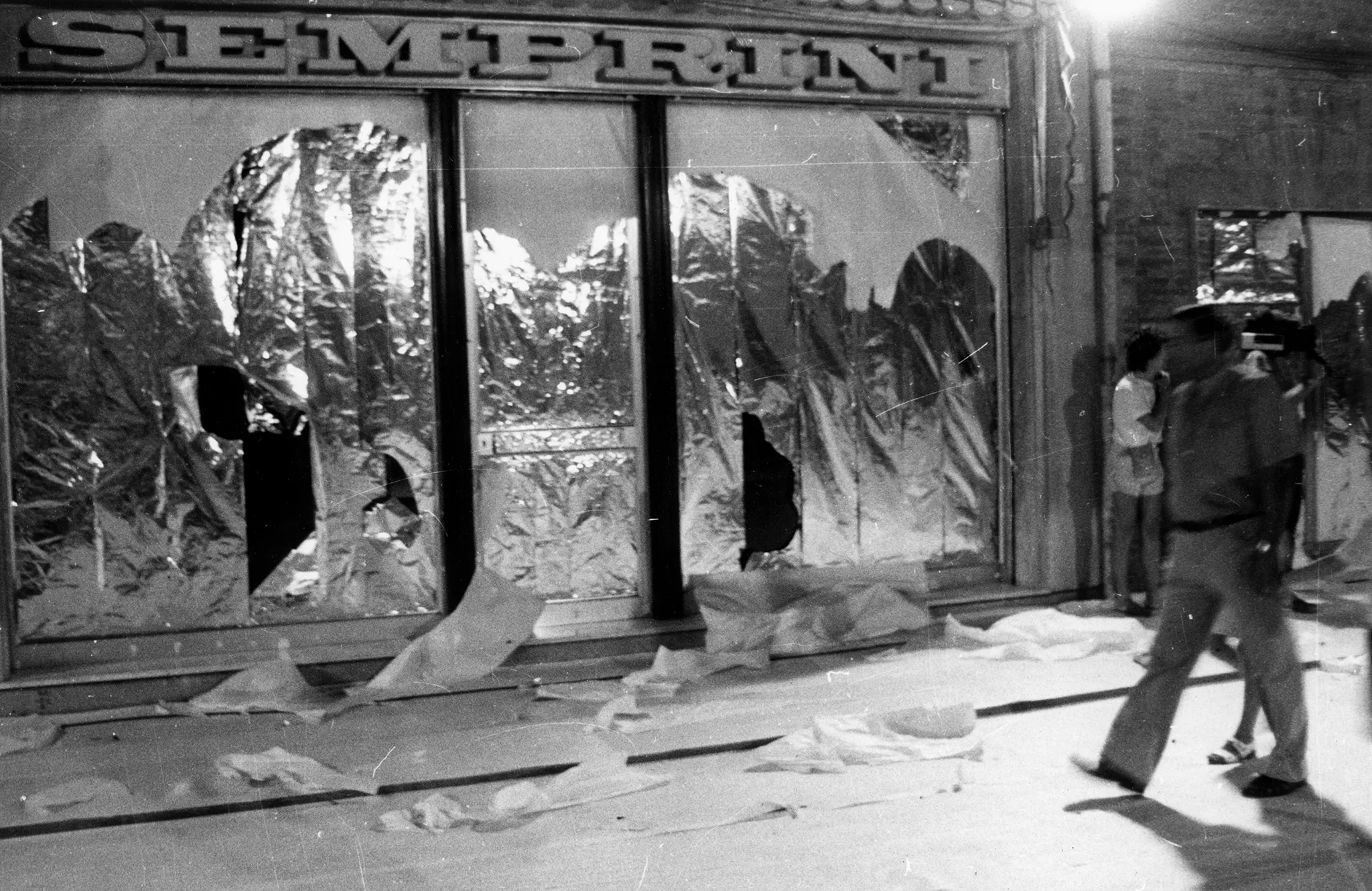 Przestrzeń miejska. Ulica wyglądająca jak tuż po zamieszkach. Pod zasłoniętymi witrynami sklepowymi leżą białe flagi. Nad drzwiami budynki widnieje napis "SEMPRINI". Z prawej strony idzie przechodzień.