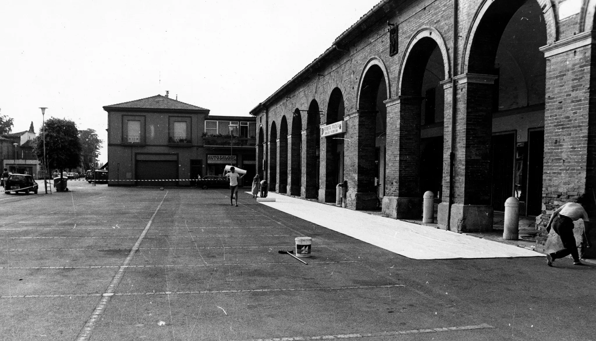 Przestrzeń miejska. Grupa aktorów i aktorek rozwija na ulicy między budynkami wielką biała płachtę. W tle widać samochody i przechodniów.