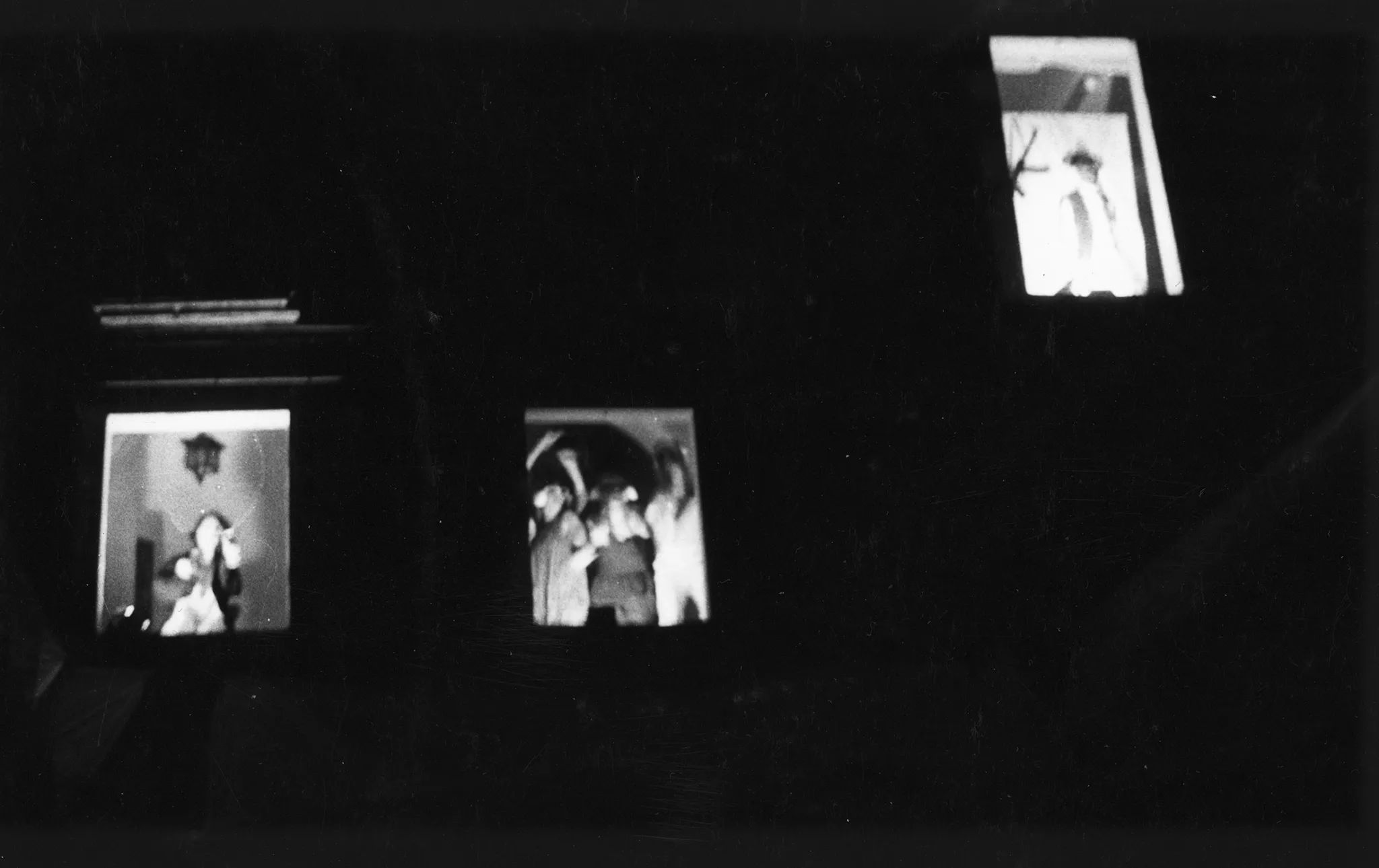 Niewyraźne zdjęcie. Trzy okna budynku - dwa niżej, jedno wyżej. W oknach stoją aktorzy.