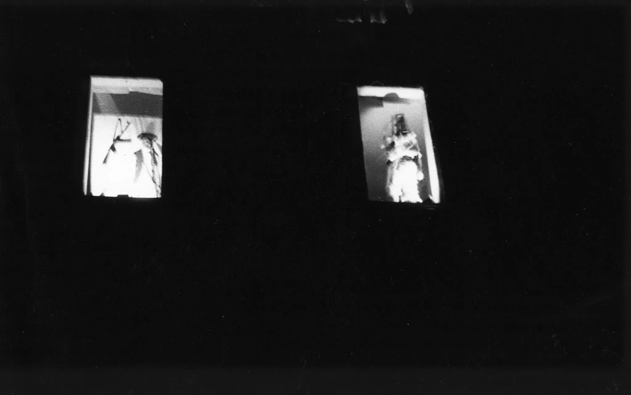 Niewyraźne zdjęcie. Dwa okna zaciemnionego budynku. W każdym oknie stoi jeden aktor zwrócony twarzą w stronę widza.