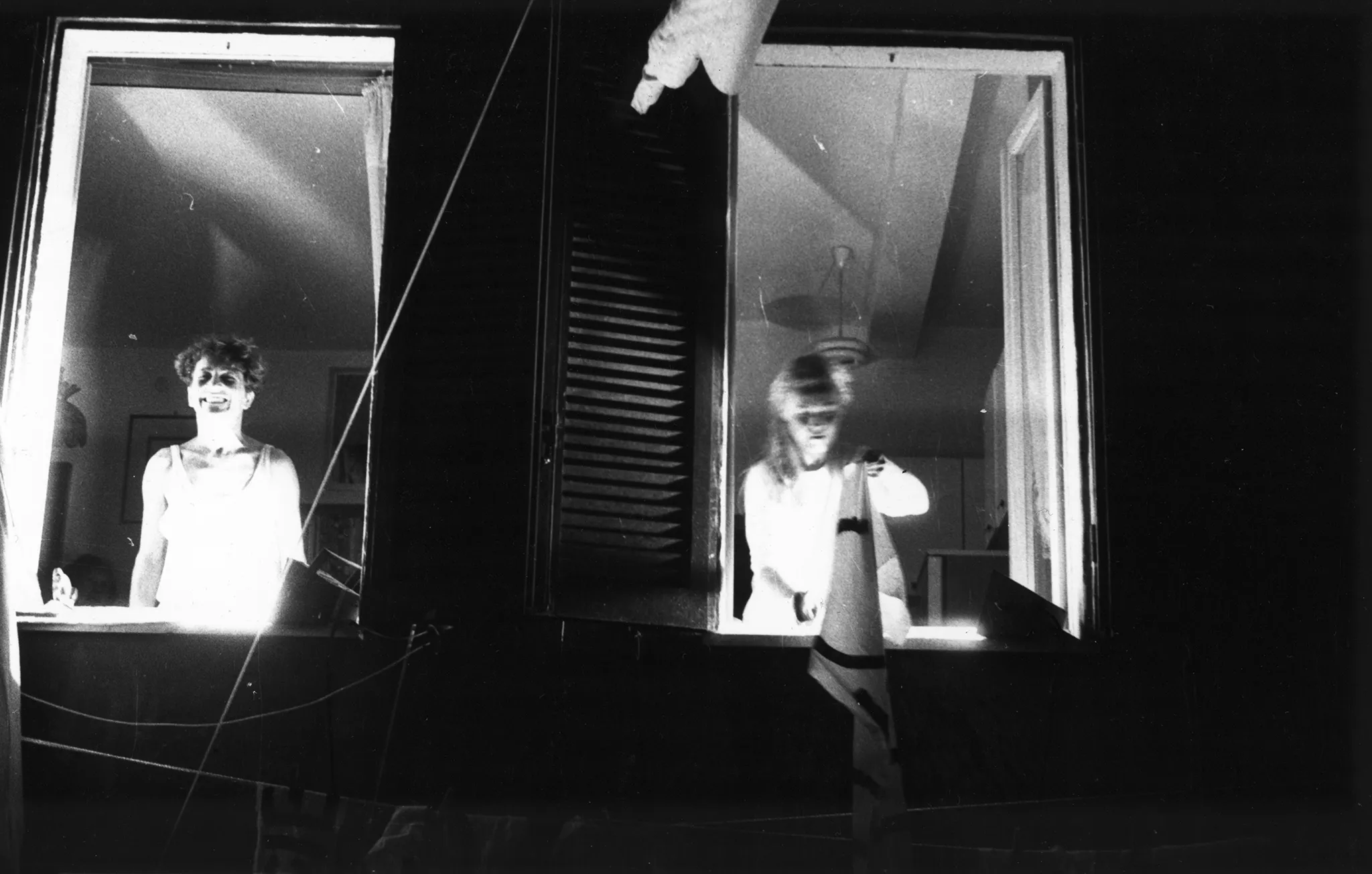 Dwa sąsiadujące okna budynku. W oknie z lewej stronie stoi aktor zwrócony twarzą w stronę widza. Ma na sobie białą koszulkę ramiączka. Uśmiecha się. Wygląda, jakby pozował do zdjęcia. W oknie z prawej strony stoi aktorka zwrócona twarzą w stronę widza. Trzyma w rękach biało-czarną chustę.