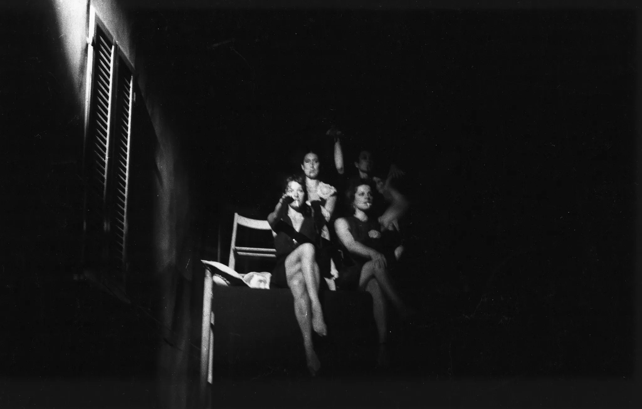 Grupa aktorek zwróconych twarzami w stronę widza siedzi na podeście. Ubrane są w czarne sukienki. Każda zakłada nogę na nogę i podnosi rękę. Palą papierosy. Po lewej stronie widać okno budynku.