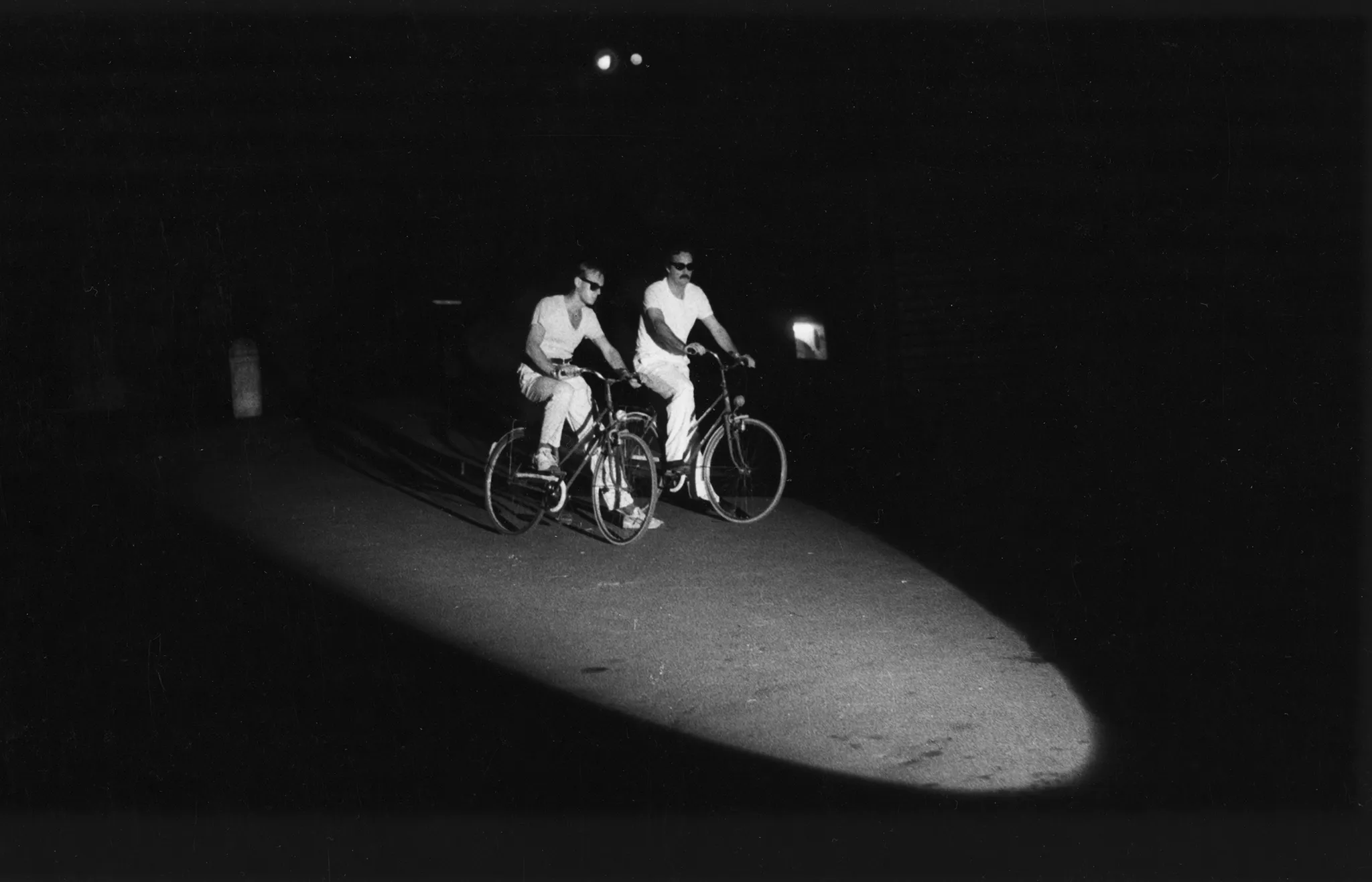 Dwóch aktorów jedzie na rowerach. Obaj ubrani są w białe spodnie i białe podkoszulki. Mają na sobie ciemne okulary przeciwsłoneczne. Oświetla ich światło z reflektora.