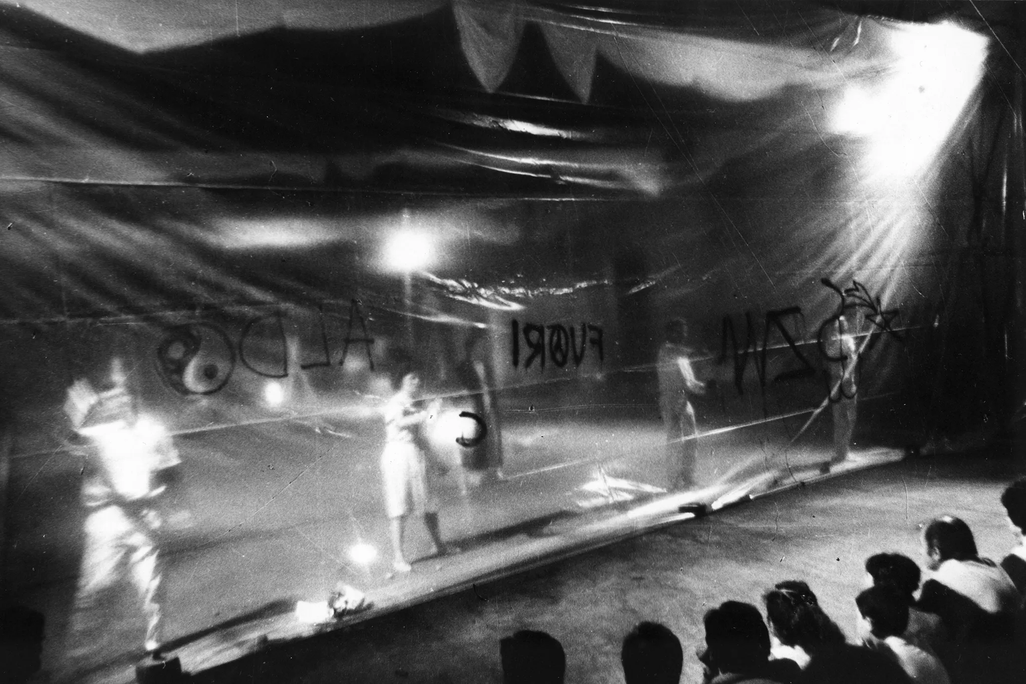 Grupa aktorów i aktorek zwrócona przodem w stronę widza stoi za przeźroczystą płachtą. Aktorzy i aktorki malują na płachcie napisy czarną farbą. Na pierwszym planie siedzi odwrócona plecami w stronę widza pubczliność oglądająca działania aktorów.