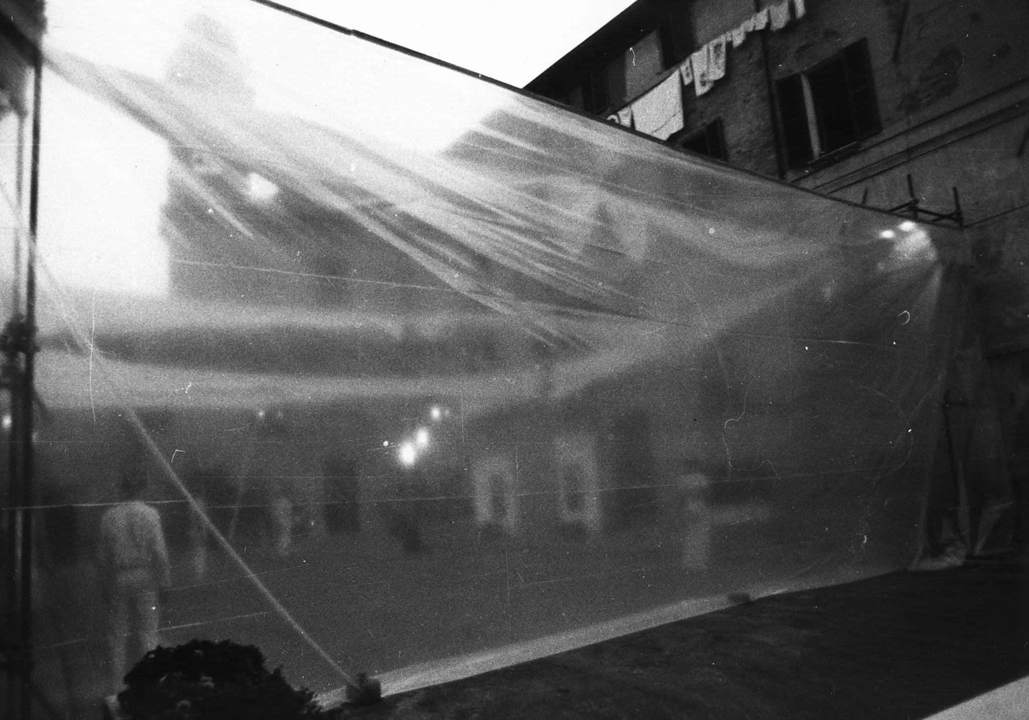 Przestrzeń miejska. Między budynkami wisi duża przeźroczysta płachta. Za płachtą stoi ledwo widoczna grupa aktorów i aktorek.