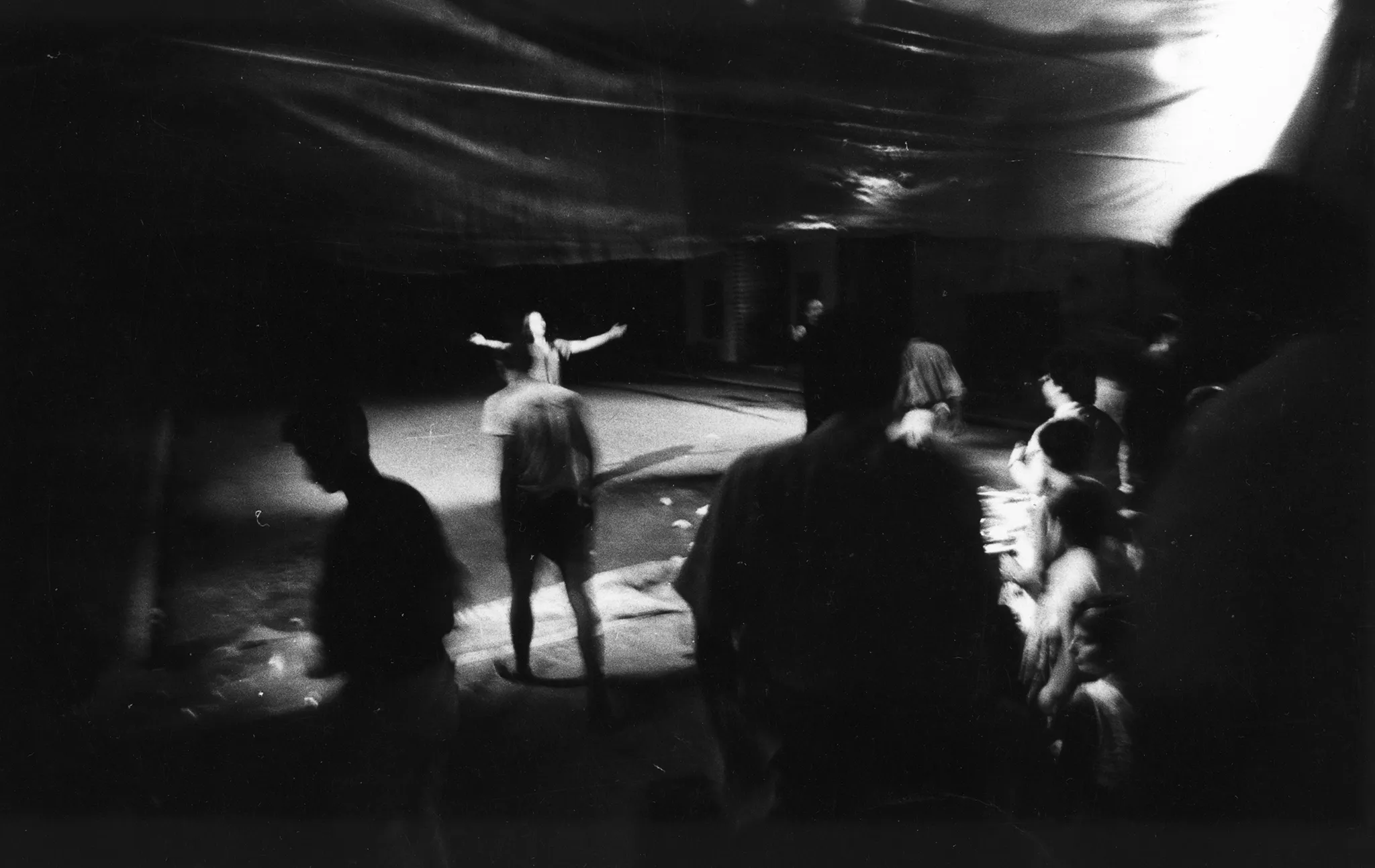 Niewyraźne zdjęcie. Przestrzeń miejska. Między budynkami, nad ulicą, wisi przeźroczysta płachta. Pod płachtą stoi zwrócona przodem w stronę widza aktorka. Wygląda, jakby śpiewała. Obok niej przechodzi grupa aktorów i aktorek. Aktorom przygląda się duża grupa widzów stojących na pierwszym planie.