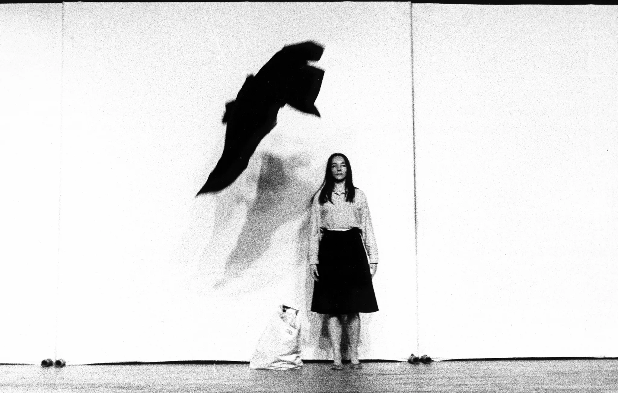 Pod białą ścianą stoi zwrócona twarzą w stronę widza aktorka. Ubrana jest w jasną koszulę i czarną spódnicę. Nad jej głową wisi czarny materiał przypominający dużego ptaka. Obok niej na podłodze stoi biały worek.