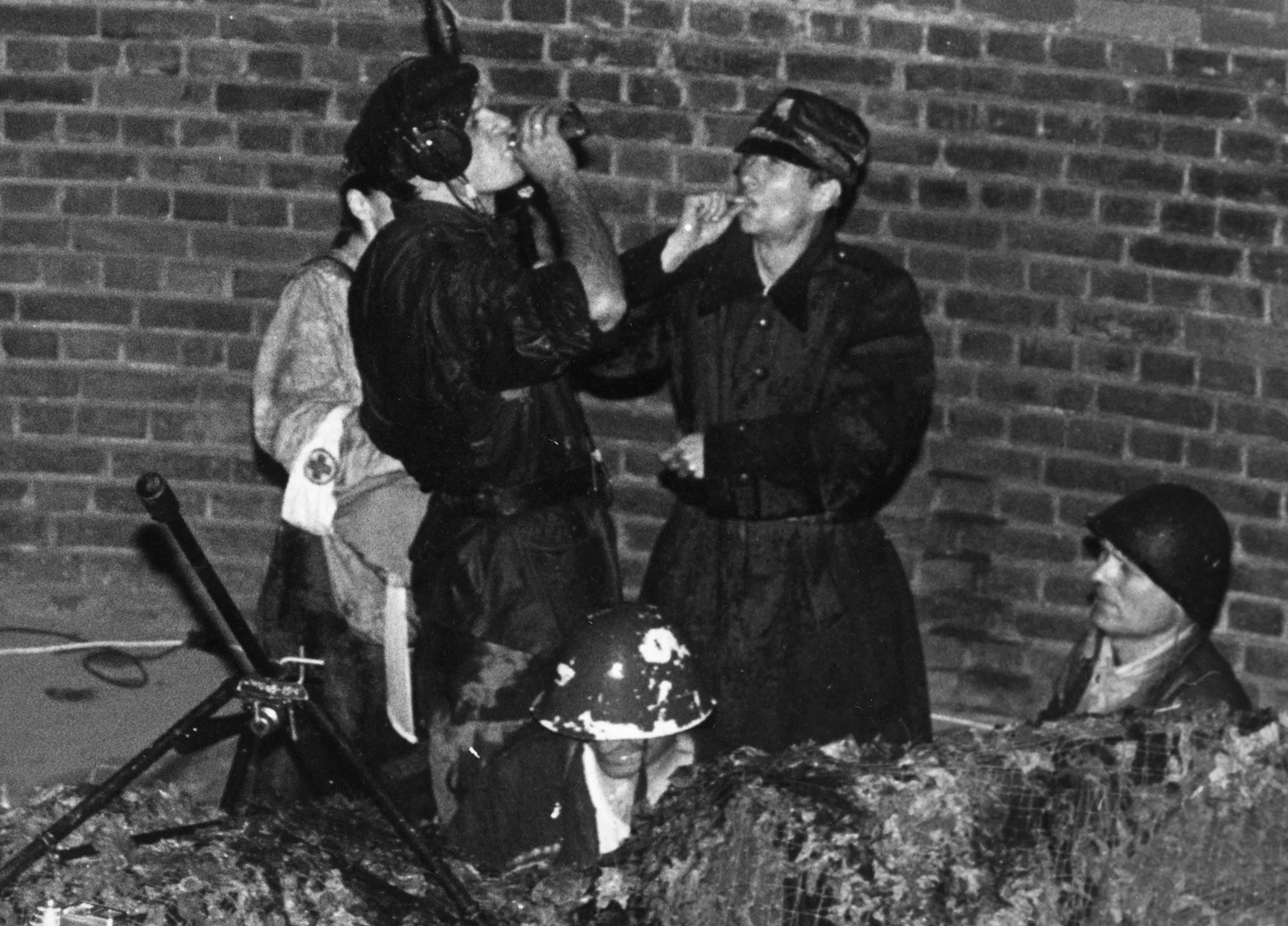 Grupa aktorów w okopie postawionym pod ceglanym murem. Ubrani są w wojskowe stroje, wyglądają na żołnierzy. Dwóch aktorów z przodu siedzi, widać tylko ich głowy. Za nimi stoich dwóch aktorów. Jeden pije alkohol, drugi pali papierosa. Za ich plecami stoi ledwo widoczny aktor wyglądający na sanitariusza. Ubrany jest na biało i ma opaskę Czerwonego Krzyża.