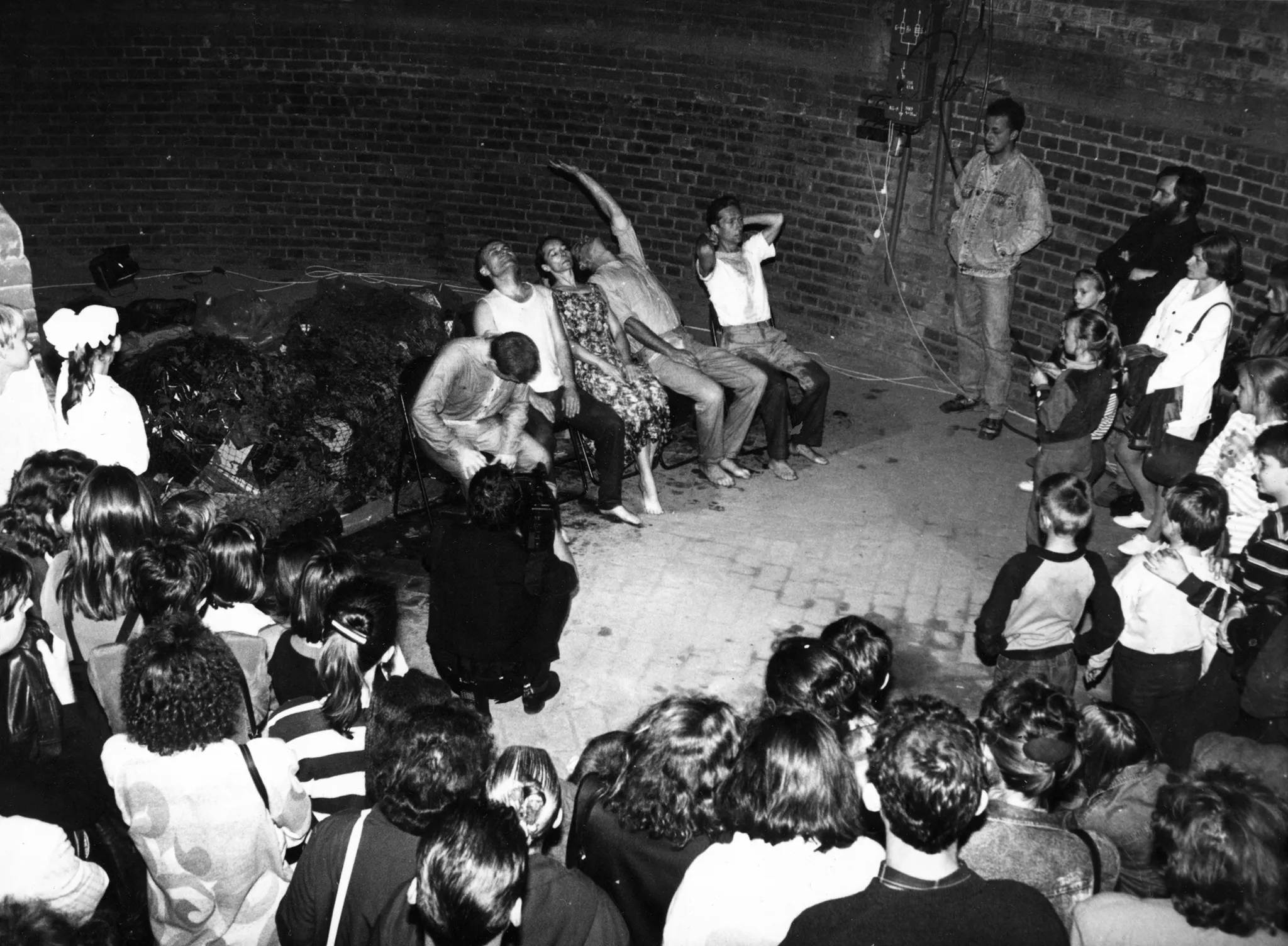 Pięcioosoba grupa aktorów i aktorek siedzi w szeregu pod murem na krzesłach. Ich ubrania wyglądają na brudne. Są boso. Tuż przy aktorach kuca kamerzysta ubrany na czarno. Przed aktorami stoi duża grupa widzów w różnym wieku. Są wśród nich dzieci i dorośli.