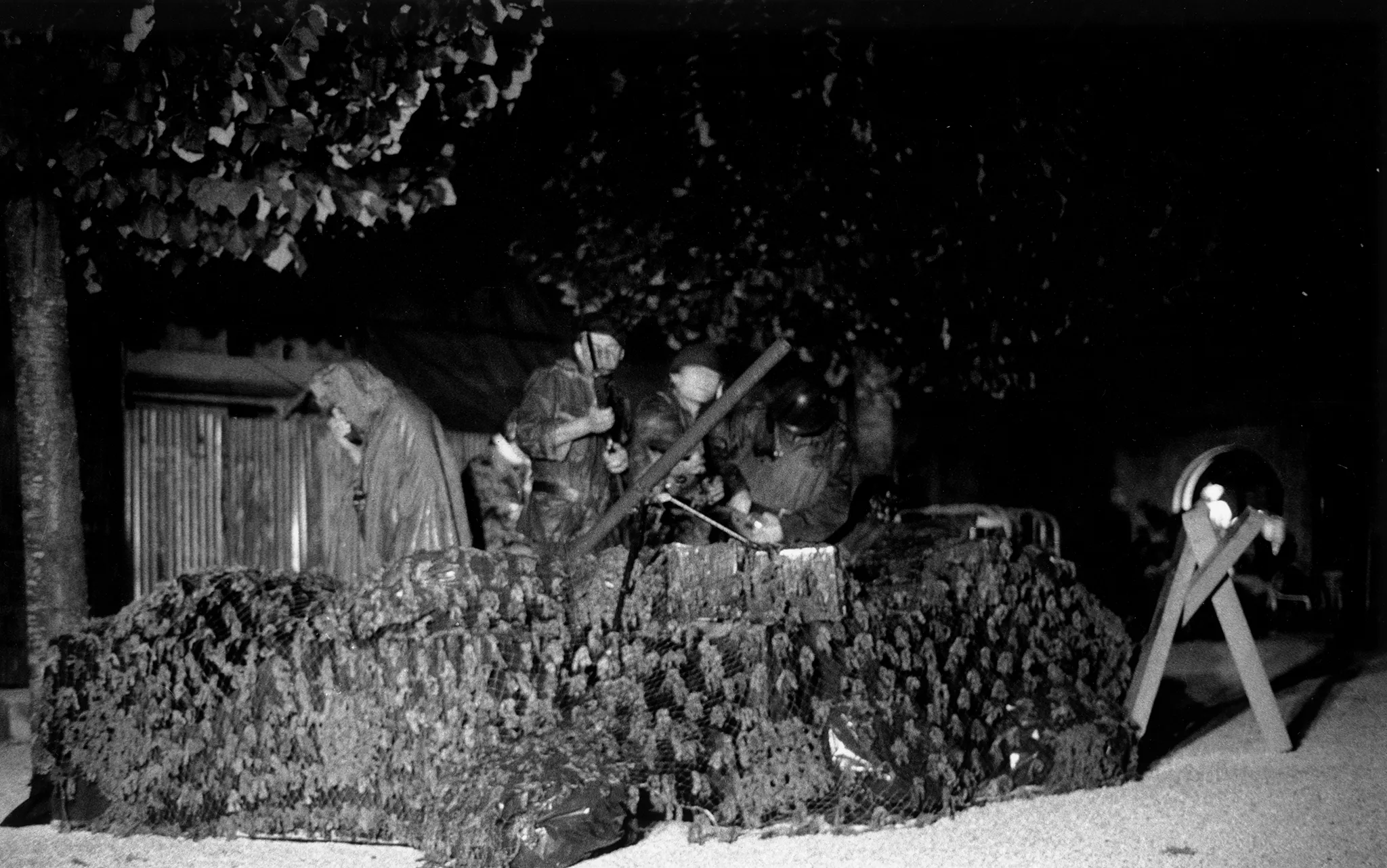 Grupa aktorów przebranych za żołnierzy stoi w okopie przodem w stronę widza. Wyglądają, jakby strzelali z karabinów. W tle widać drzewa i budynek z blachy.