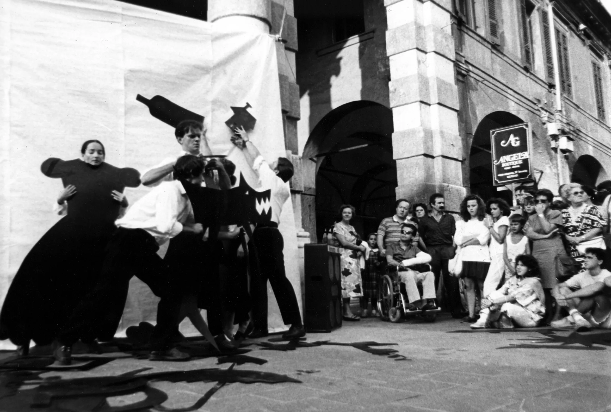 Przestrzeń miejska. Pod białą płachtą zawieszoną na kolumny budynku stoi grupa aktorów i aktorek. W rękach trzymają dwuwymiarowe czarne rekwizyty wyglądające na wycięte z papieru. Stojąca smotnie aktorka przymierza dwuwymiarową suknię. Grupa obok niej trzyma takie rekwizyty, jak butelka, domek i wilk. Po prawej stronie stoją widzowie oglądający aktorów. Za ich plecami stoi budynek.