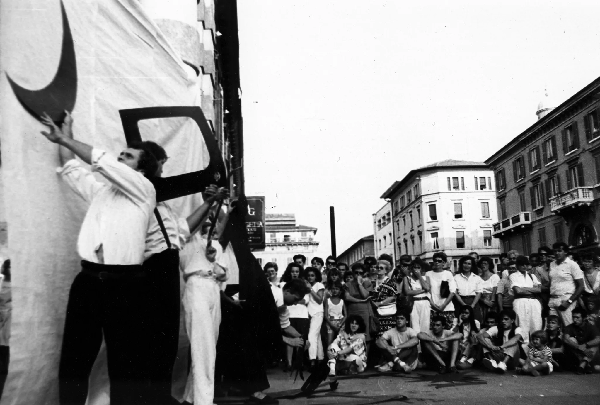 Przestrzeń miejska. Po lewej stronie pod białą płachtą grupa aktorów i aktorek trzyma w rękach trudne do zidentyfikowania czarne dwuwymiarowe rekwiyty. Rekwizyty wyglądają na wycięte z papieru. Po prawej stronie stoi duża grupa widzów oglądających aktorów. W tle widać budynki.