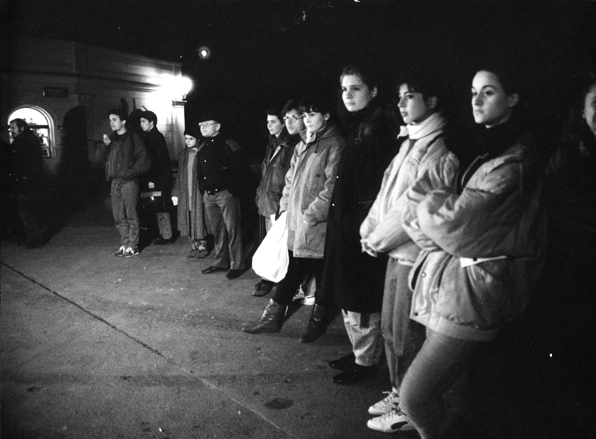 Przestrzeń miejska. Publiczność zwrócona profilem w stronę widza. Najwyraźniej coś ogląda. Wszyscy ubrani są w ciepłe ubrania, takie jak spodnie jeansowe i kurtki. W tle widac oświetlony niewielki budynek.