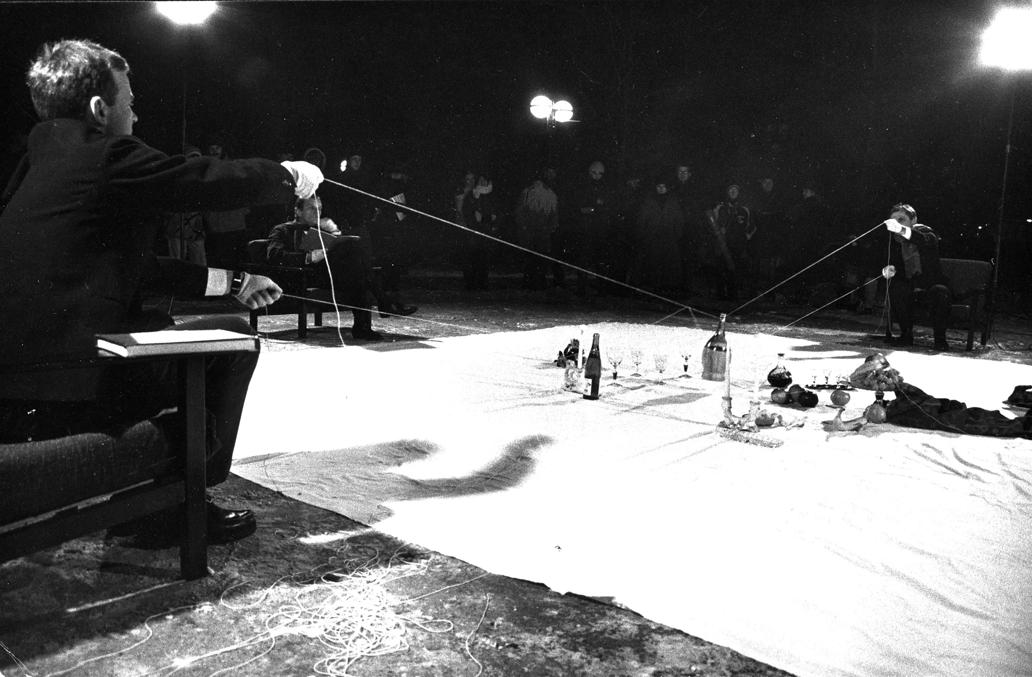 Po dwóch przeciwnych stronach białej kwadratowej płachty siedzą na krzesłach dwaj aktorzy. Obaj trzymają linki, na których zawieszone są przedmioty, takie jak butelki, kieliszki i świece. Po lewej stronie na krześle siedzi aktor, który czyta książkę. W tle widać niewielką grupę widzów, którzy przyglądają się aktorom.