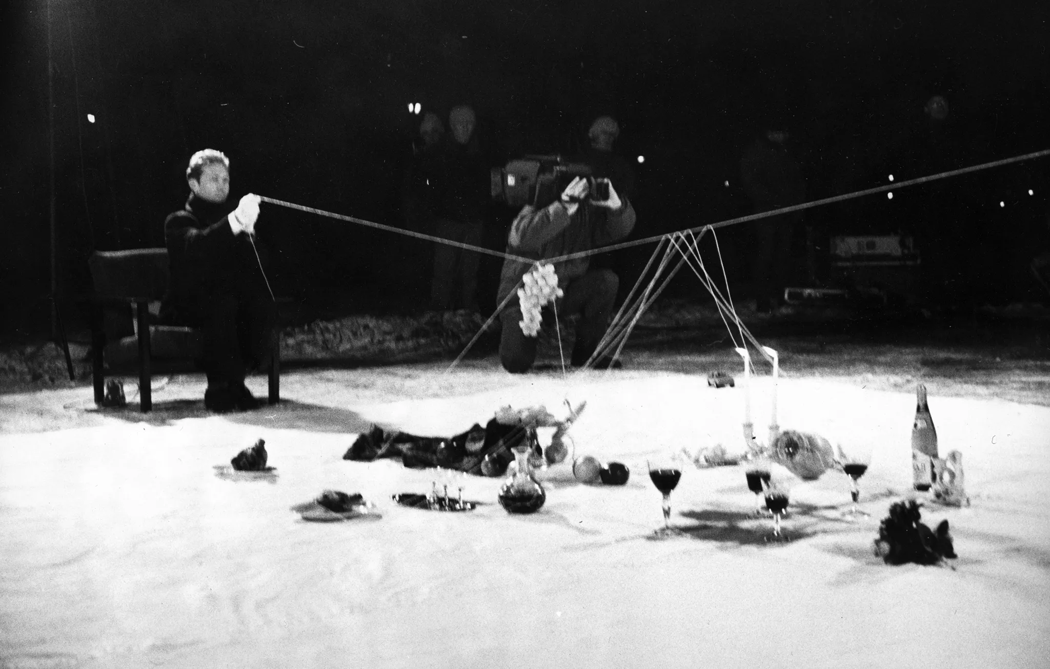 Na białej łachcie leżą przedmioty, takie jak butelki, kieliszki, świece, kwiaty i owoce. Przy płachcie na krześle siedzi ubrany na czarno aktor trzymający w ręce napiętą linkę. Na lince wiszą winogrona. W rogu białej płachty klęczy kamerzysta z twarzą zasłoniętą przez kamerę. W tle stoi mała grupa widzów.