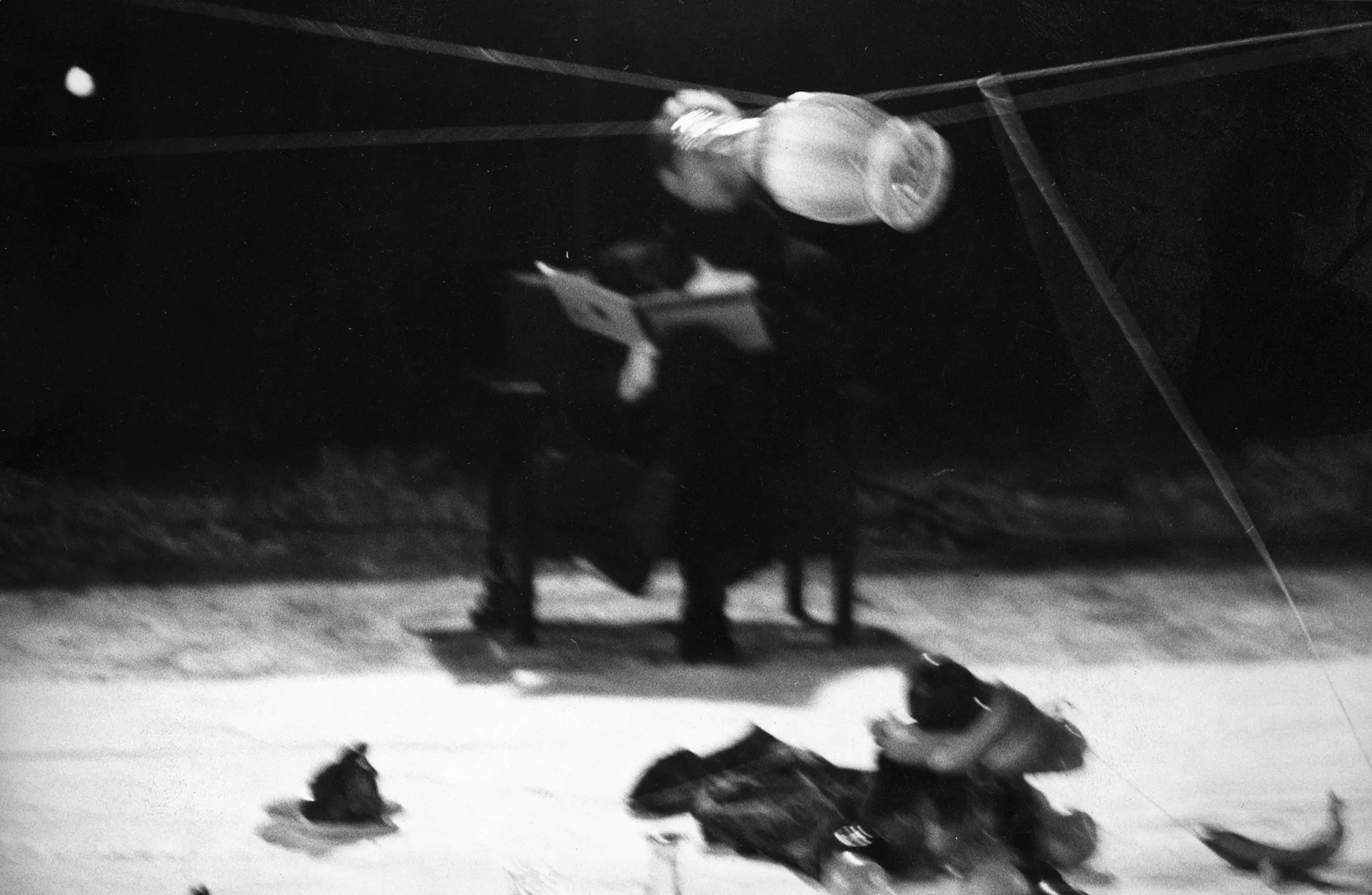 Niewyraźne zdjęcie. Na białej płachcie leżą trudne do zidentyfikowania przedmioty. Nad nimi na lince wisi pusta butelka. W tle na krześle siedzi aktor zwrócony przodem w stronę widza. Na kolanach trzyma książkę.