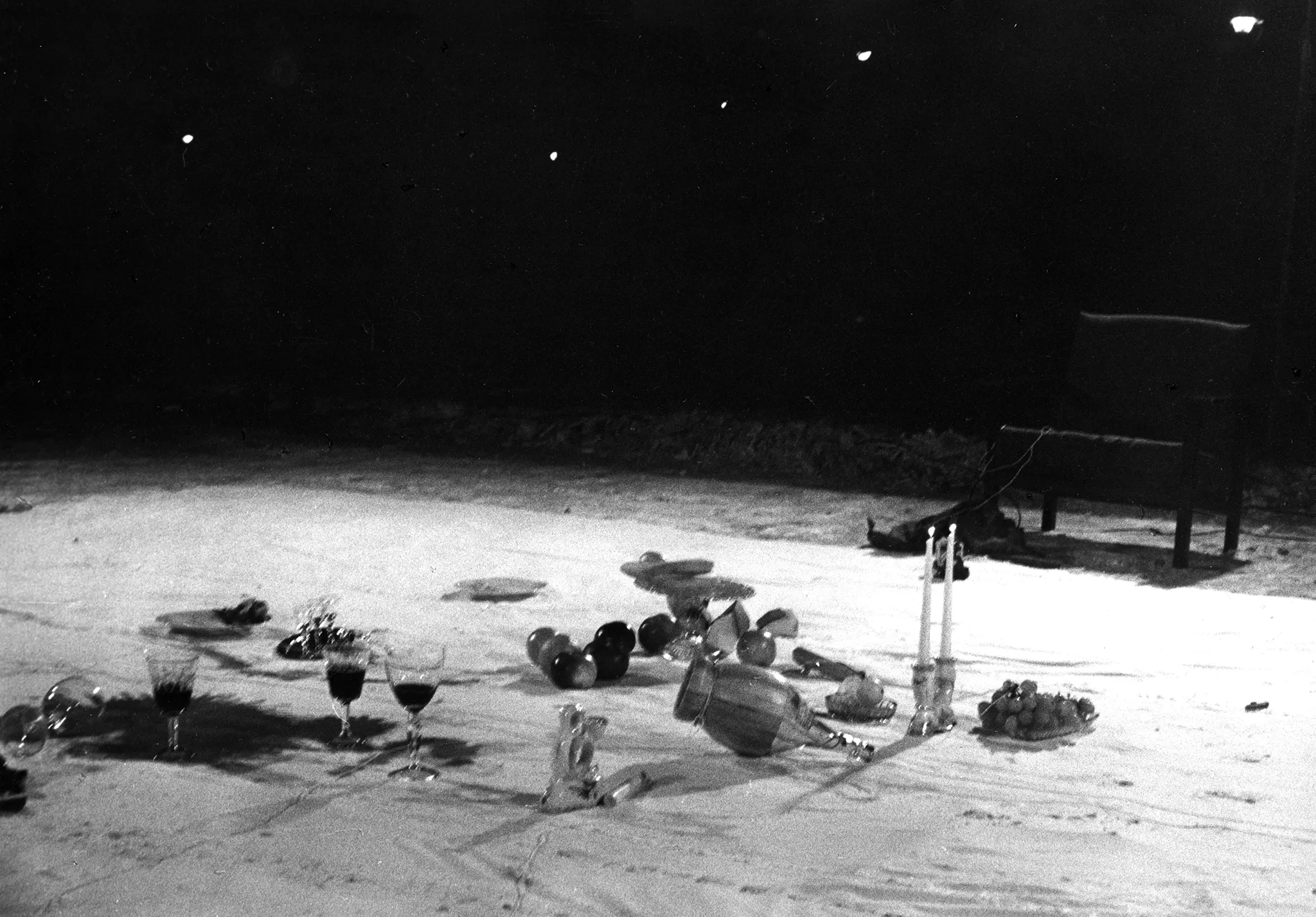 Na białej płachcie leżą przedmioty, takie jak kieliszki, świece i owoce. Niektóre przedmioty są przewrócone. Po lewej stronie płachta jest brudna od wina. Obok białej płachty stoi puste krzesło.