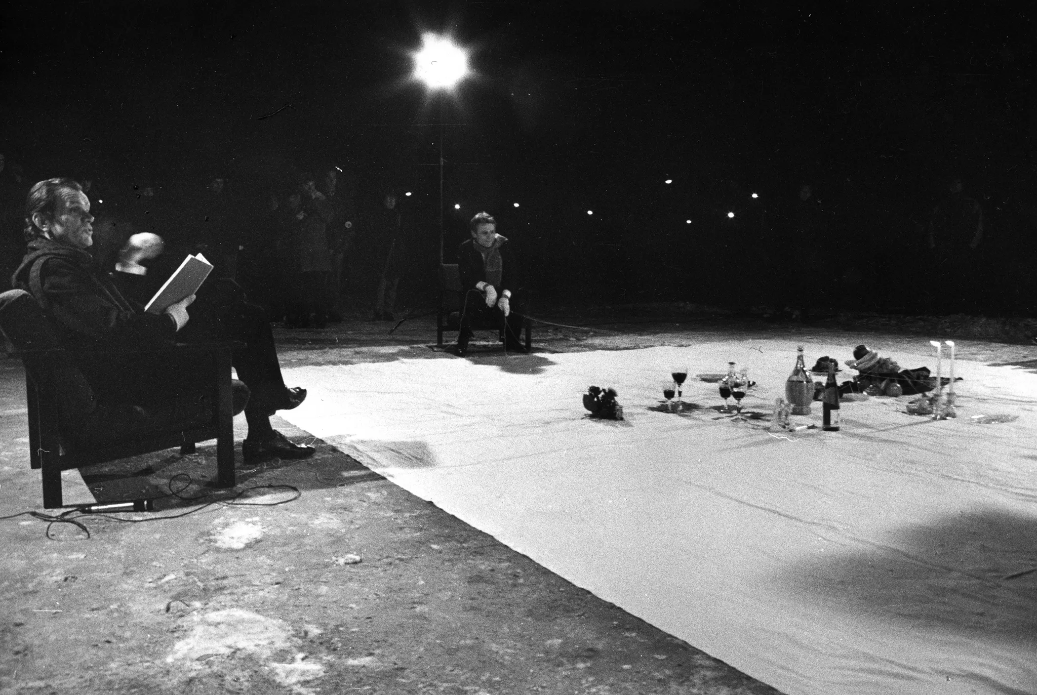 Na białej płachcie leżą przedmioty, takie jak kieliszki, świece, owoce i butelki. Po lewej stronie płachty na krześle siedzi aktor. Ubrany jest na czarno. W rękach trzyma książkę i coś mówi. Z tyłu płachty na krześle siedzi aktor trzymający w rękach napiętą linkę. Za jego plecami stoi reflektor i grupa widzów.