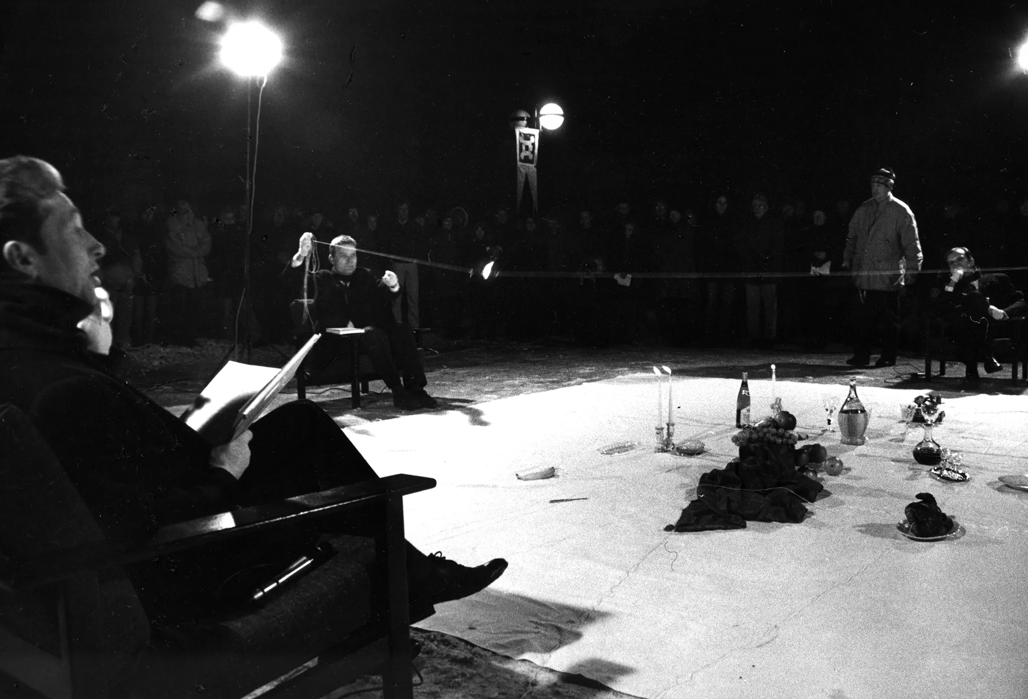 Na białej płachcie leżą przedmioty, takie jak kieliszki, świece, owoce i butelki. Po lewej stronie płachty na krześle siedzi aktor. Ubrany jest na czarno. W rękach trzyma książkę i mówi coś do aktora siedzącego na krześla po drugiej stronie płachty. Z tyłu płachty na krześle siedzi aktor trzymający w rękach napiętą linkę. Za jego plecami stoi reflektor i grupa widzów.