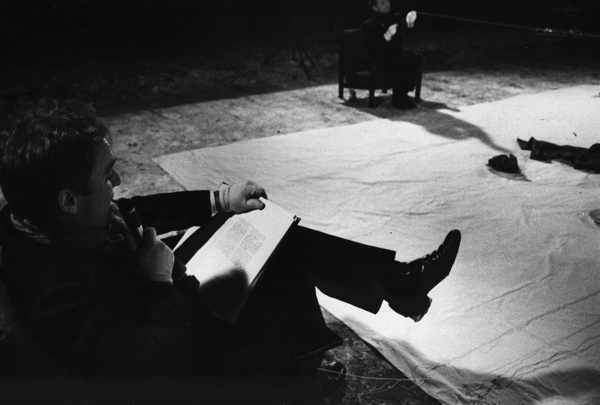 Po lewej stronie białej płachty na krześle siedzi aktor. Ubrany jest na czarno. Na kolanach trzyma książkę i mówi coś do mikrofonu. Z tyłu płachty na krześle siedzi aktor trzymający w rękach napiętą linkę.