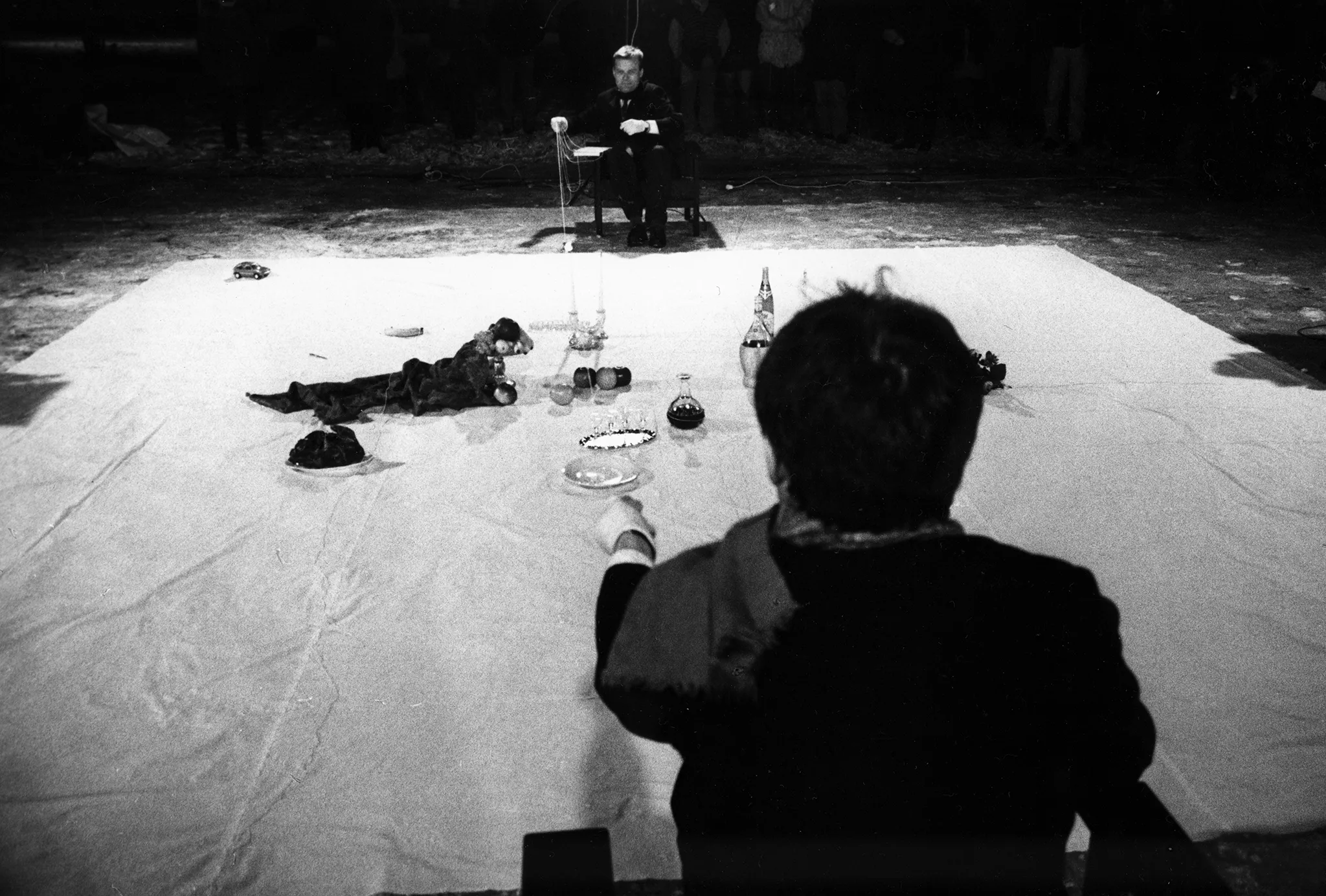 Na białej płachie leżą przedmioty, takie jak owoce, butelki i talerzyki. Po dwóch przeciwnych stronach płachty siedzi dwóch aktorów - jeden zwrócony plecami, drugi twarzą w stronę widza. Obaj trzymają w rękach napiętą linkę.