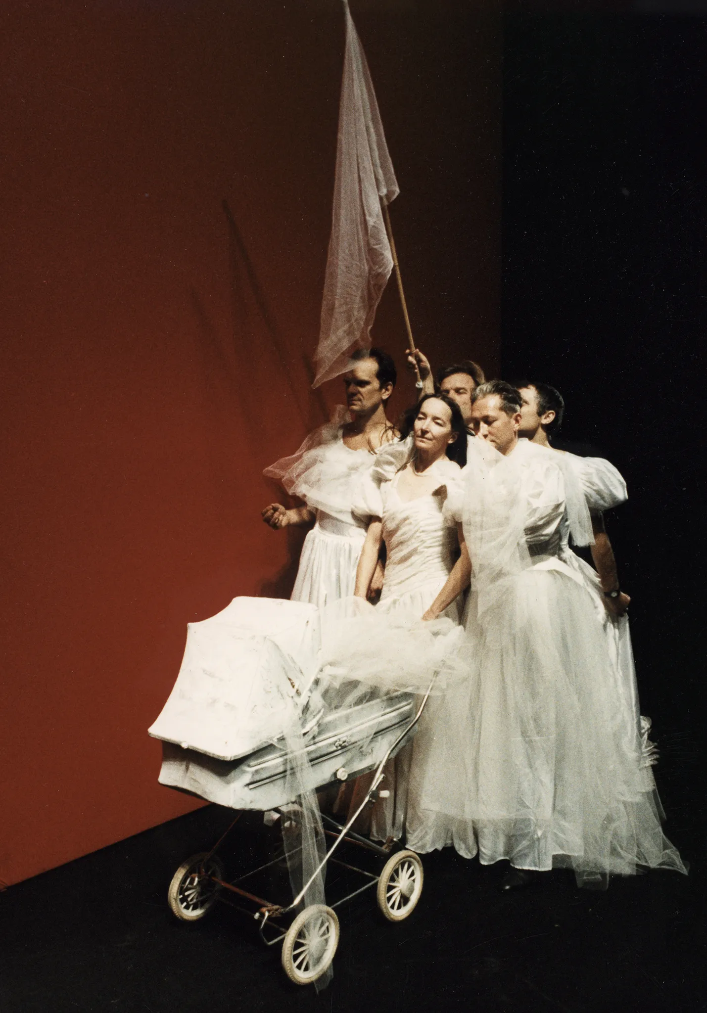 Na tle czerwonej ściany przez scenę przechodzi czterech aktorów i jedna aktorka. Idą w zwartej grupie. Wszyscy ubrani są w białe suknie ślubne. Aktorka idzie z przodu i pcha biały wózek dziecięcy. Aktor idący z tyłu trzyma w ręce białą flagę na kiju.