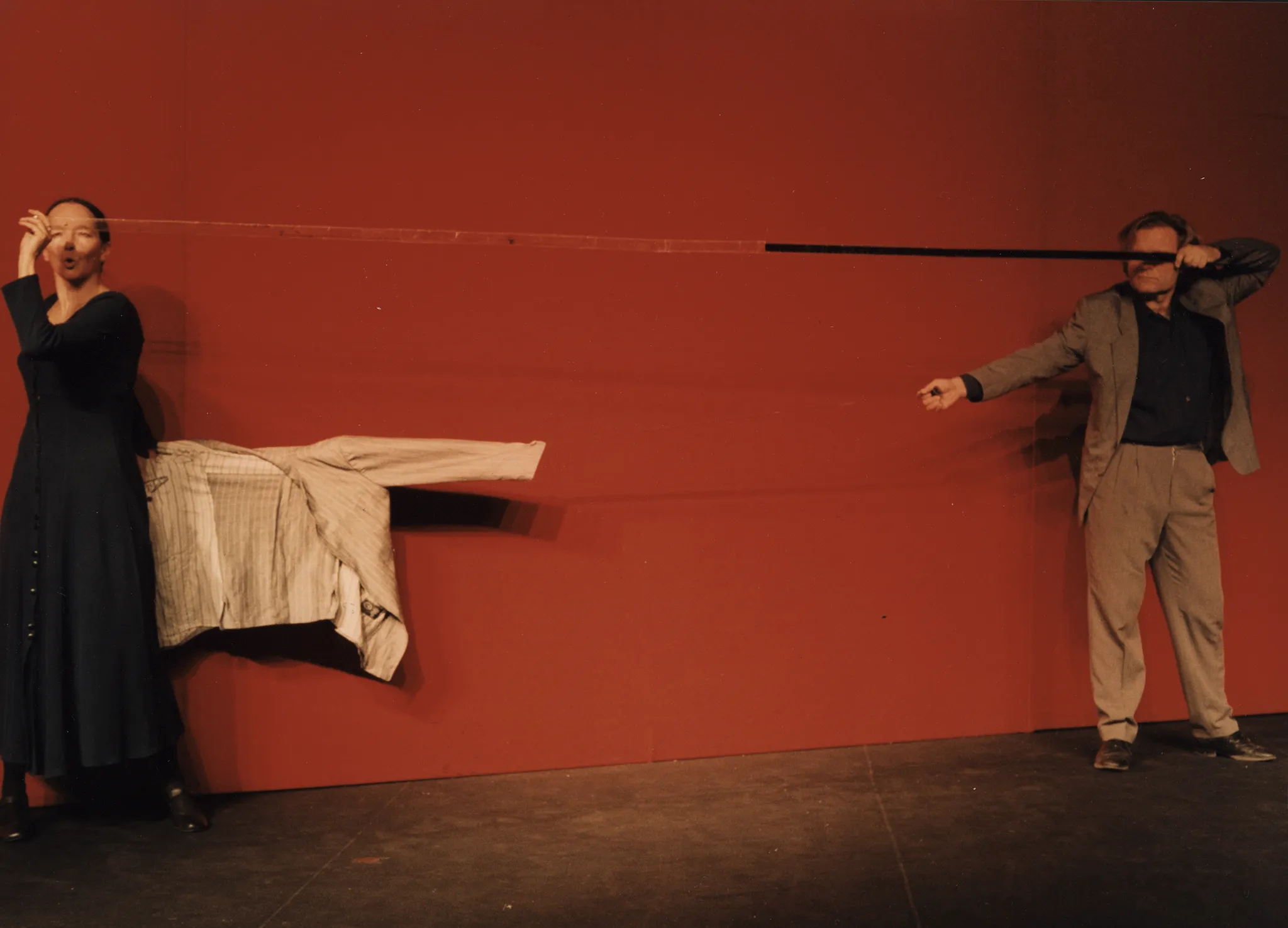 Na tle czerwonej ściany stoi aktorka i aktor. Aktorka stoi z lewej strony, aktor z prawej. Trzymają w rękach długą taśmę, z lewej strony przeźroczystą, z prawej czarną. Aktorka ubrana jest w granatową sukienkę. Za nią na lince wisi biała marynarka. Aktor ubrany jest w szary garnitur i granatową koszulę. Jego oczy zasłania czarny fragment taśmy.