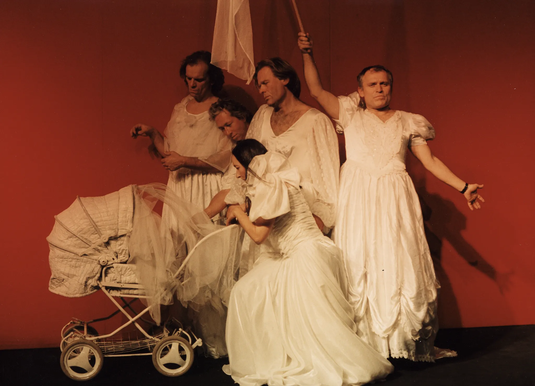 Na tle czerwonej ściany stoi czterech aktorów i jedna aktorka. Trzech aktorów i aktorka zwróceni są profilem w stronę widza. Zaglądają do białego wózka dziecięcego. Jeden aktor zwrócony jest twarzą w stronę widza. Trzyma w ręce białą flagę na kiju. Wszyscy ubrani są w białe suknie ślubne.