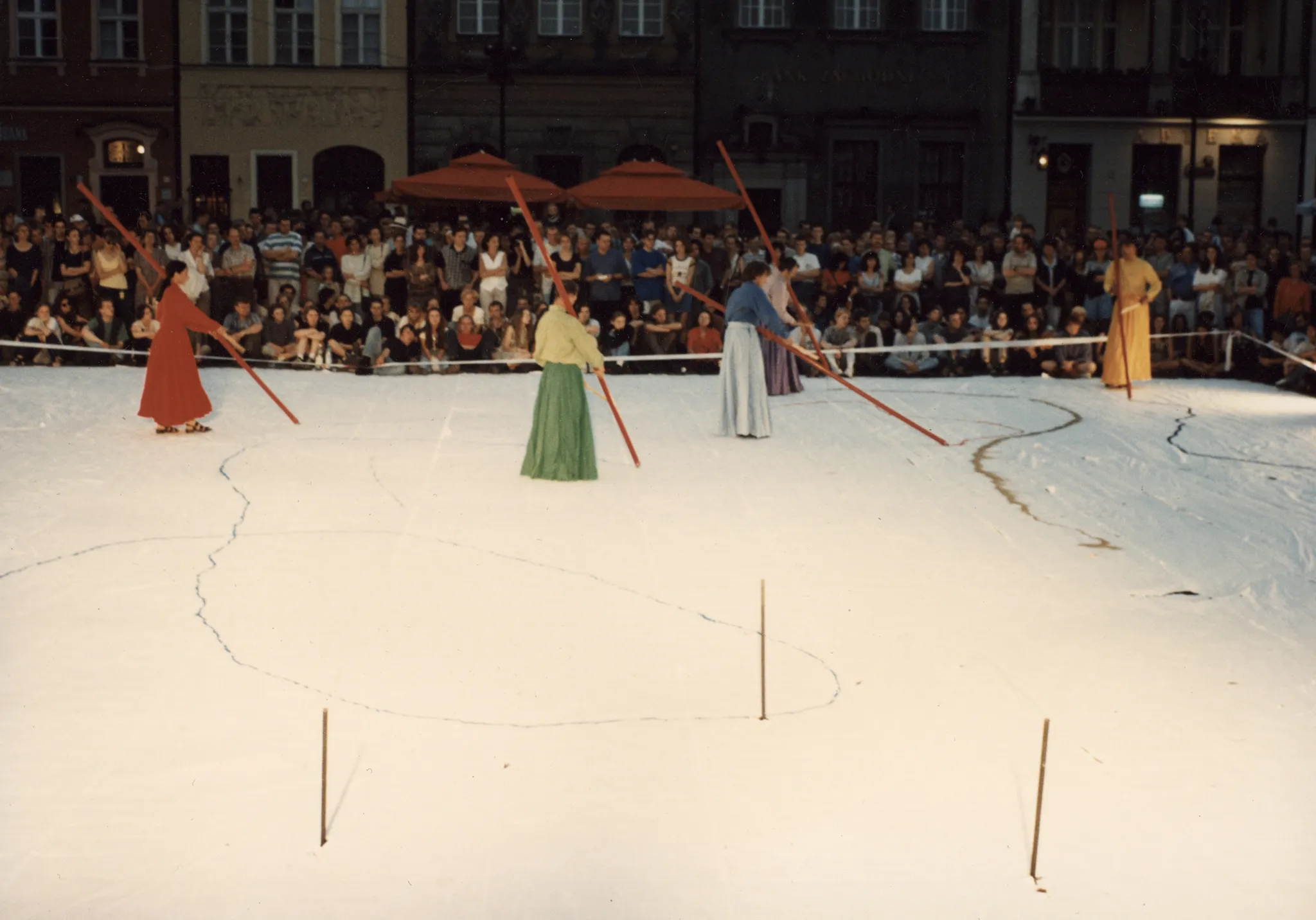 Duża biała scena ustawiona w przestrzeni miejskiej. Scena wygląda, jakby był na niej śnieg. Na scenie stoi czterosoobowa grupa aktorów i aktorek ubranych w kolorowe suknie w stylu azjatyckim. Trzymają w rękach długie czerwone kije i rysują wzory na scenie. W tle pod budynkami stoi publiczność zwrócona twarzą w stronę widza.