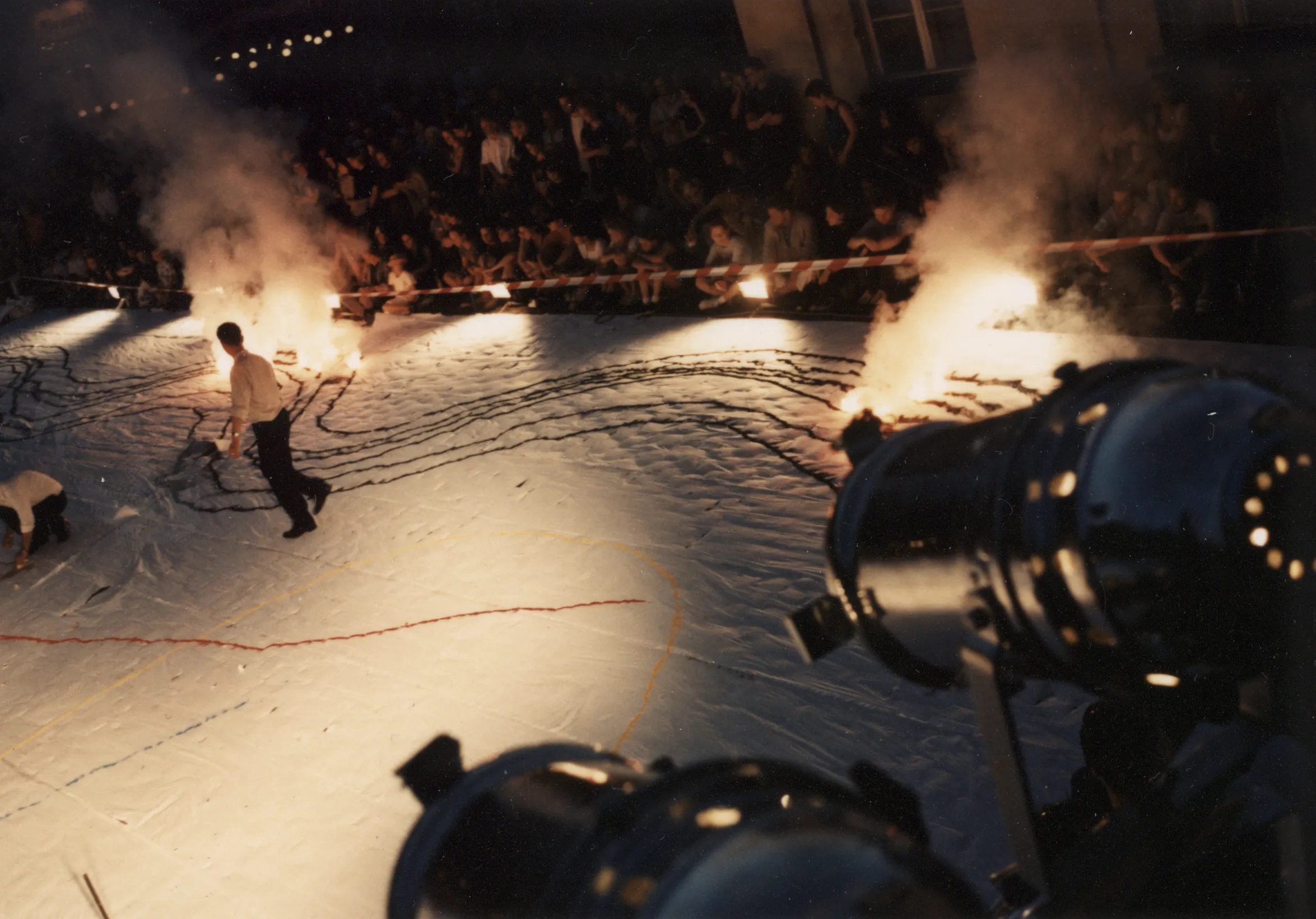 Scena ustawiona w przestrzeni miejskiej. Scena jest biała. Leżą na niej długie wzory w różnych kolorach. W niektórych miejscach są podpalone. Na scenie wśród wzorów chodzi dwóch aktorów ubranych w czarne spodnie i białe koszule. Pod sceną siedzi i stoi publiczność.