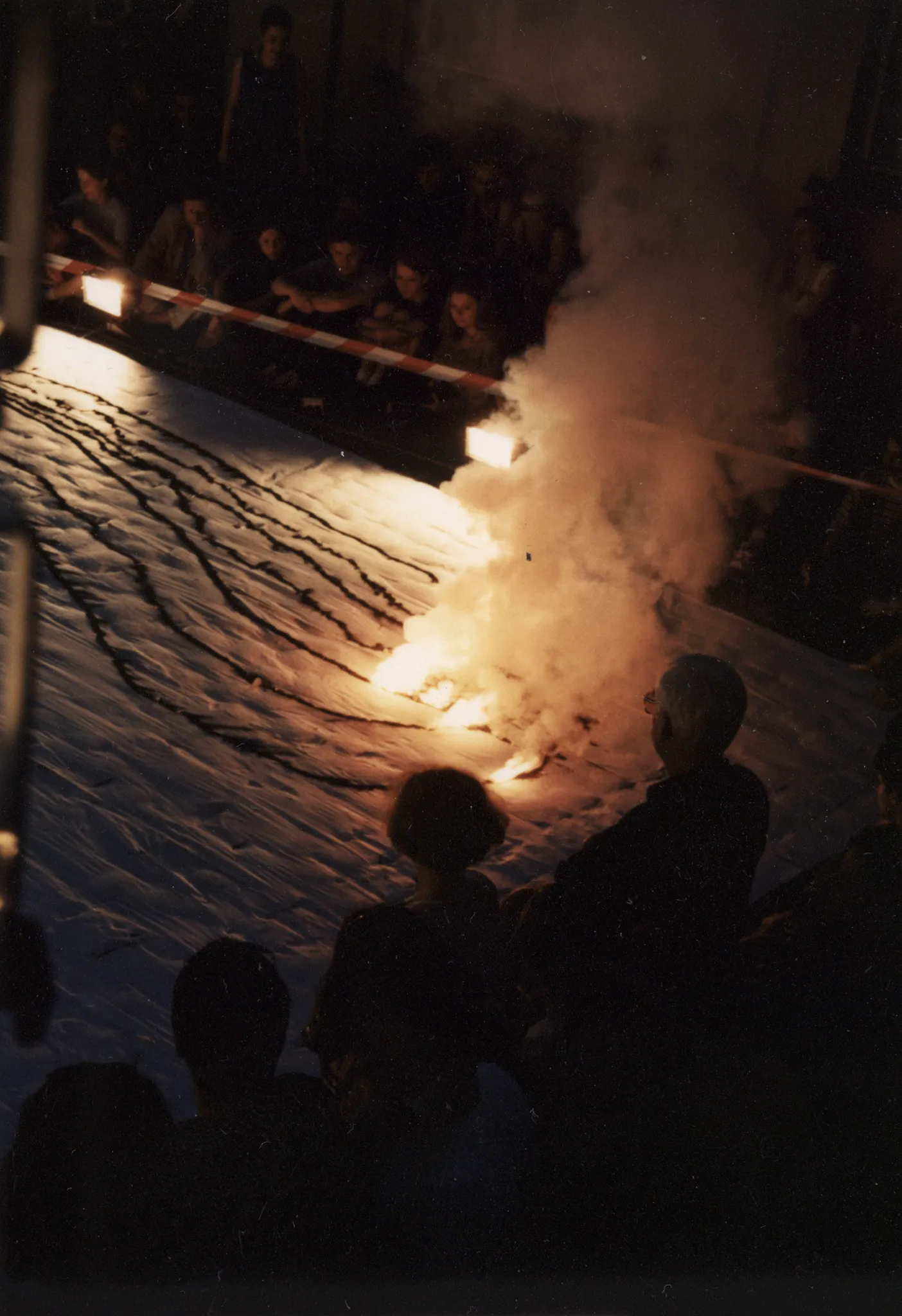 Scena ustawiona w przestrzeni miejskiej. Scena jest biała. Leżą na niej długie wzory w różnych kolorach. W niektórych miejscach są podpalone. Unosi się z nich dym. Pod sceną siedzi i stoi publiczność.