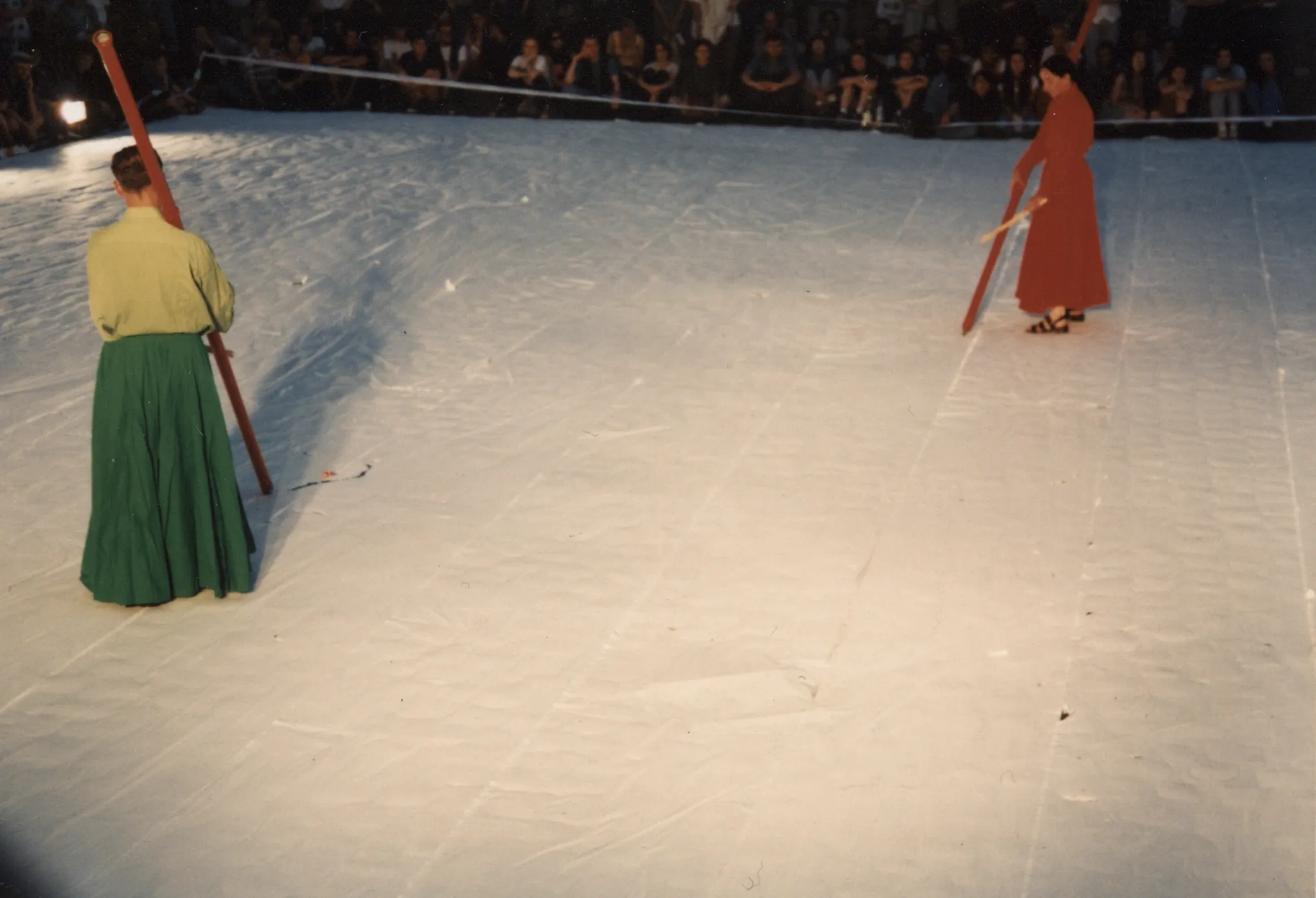 Scena ustawiona w przestrzeni miejskiej. Scena jest biała. Na scenie stoi aktorka i aktor. Aktorka ubrana jest w czerwoną suknię. Aktor ubrany jest w zieloną spódnicę i jasnozieloną koszulę. Oboje trzymają w rękach długie czerwone kije, z których wysypuje się proszek. W tle widać publiczność.