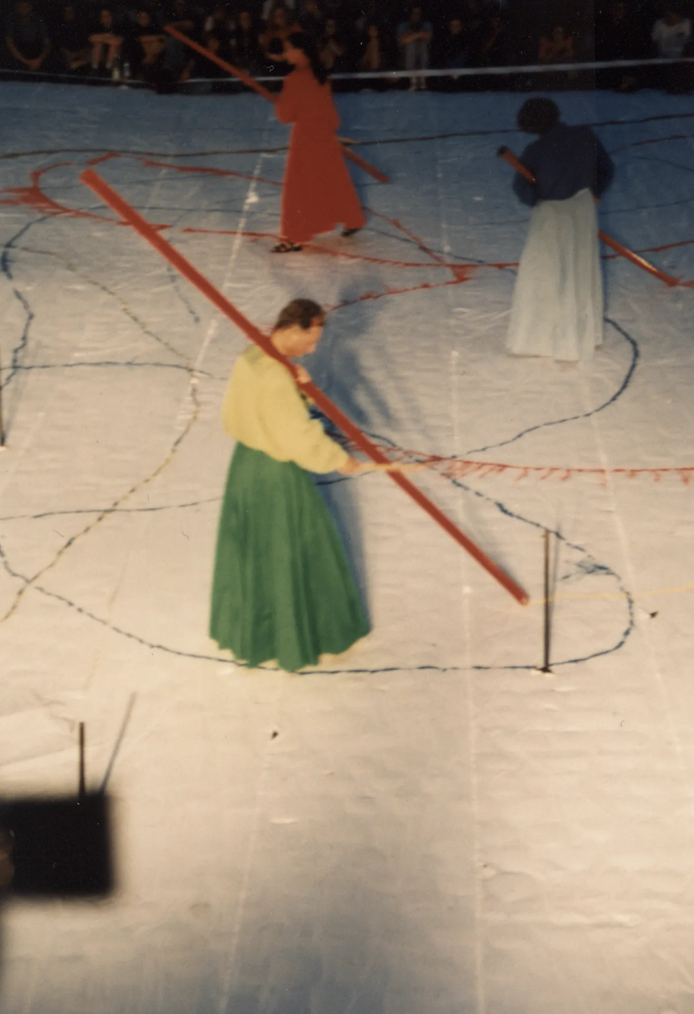 Scena ustawiona w przestrzeni miejskiej. Scena jest biała, pełna kolorowych linii. Na scenie stoi aktorka, osoba o trudnej do określenia płci i jeden aktor. Jedna z aktorkek ubrana jest w czerwoną suknię. Osoba o trudnej do określenia płci ubrana jest w białą spódnicę i granatową koszulę.  Aktor ubrany jest w zieloną spódnicę i jasnozieloną koszulę. Wszyscy trzymają w rękach długie czerwone kije, z których wysypuje się proszek. W tle widać publiczność.