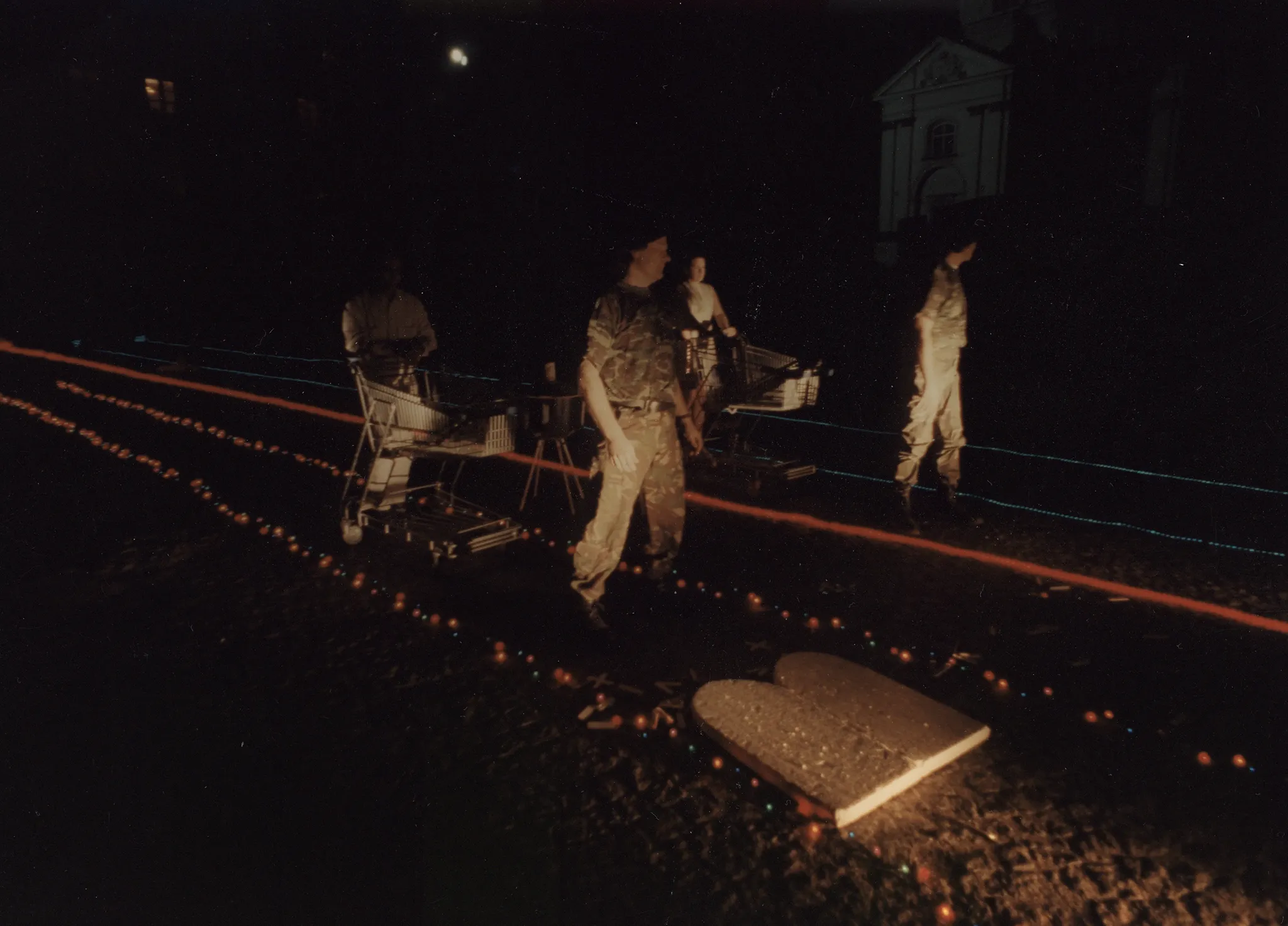 Niewyraźne zdjęcie. Noc. Przestrzeń miejska. Noc. Przestrzeń miejska. Na piasku leżą trasy wyznaczone przez czerwone i niebieskie lampki. Pośród lampek stoją aktorzy i aktorki. Wszyscy ubrani są w ubrania wojskowe. W tle widać budynki.