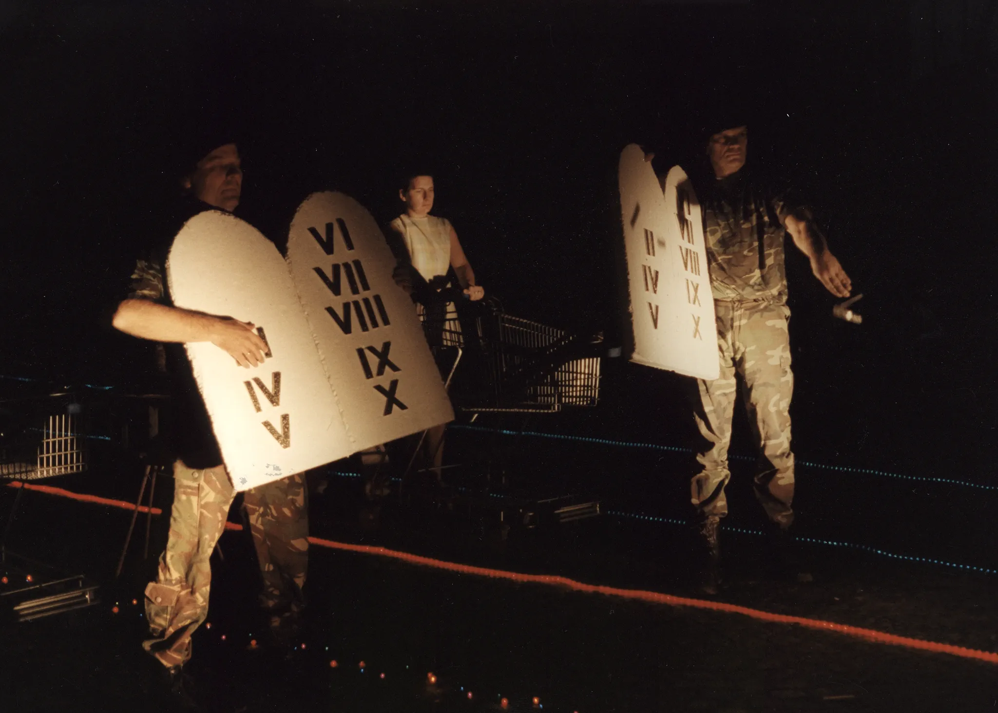 Noc. Na piasku leżą trasy wyznaczone przez czerwone i niebieskie lampki. Pośród lampek stoją aktorzy i wózki sklepowe. Dwóch aktorów w ubraniach wojskowych trzyma tablice z rzymskimi cyframi kończącymi się na "X". Tablice przypominają Dekalog. Aktorzy odrywają w tablic kolejne cyfry i rzucają je na ziemię.